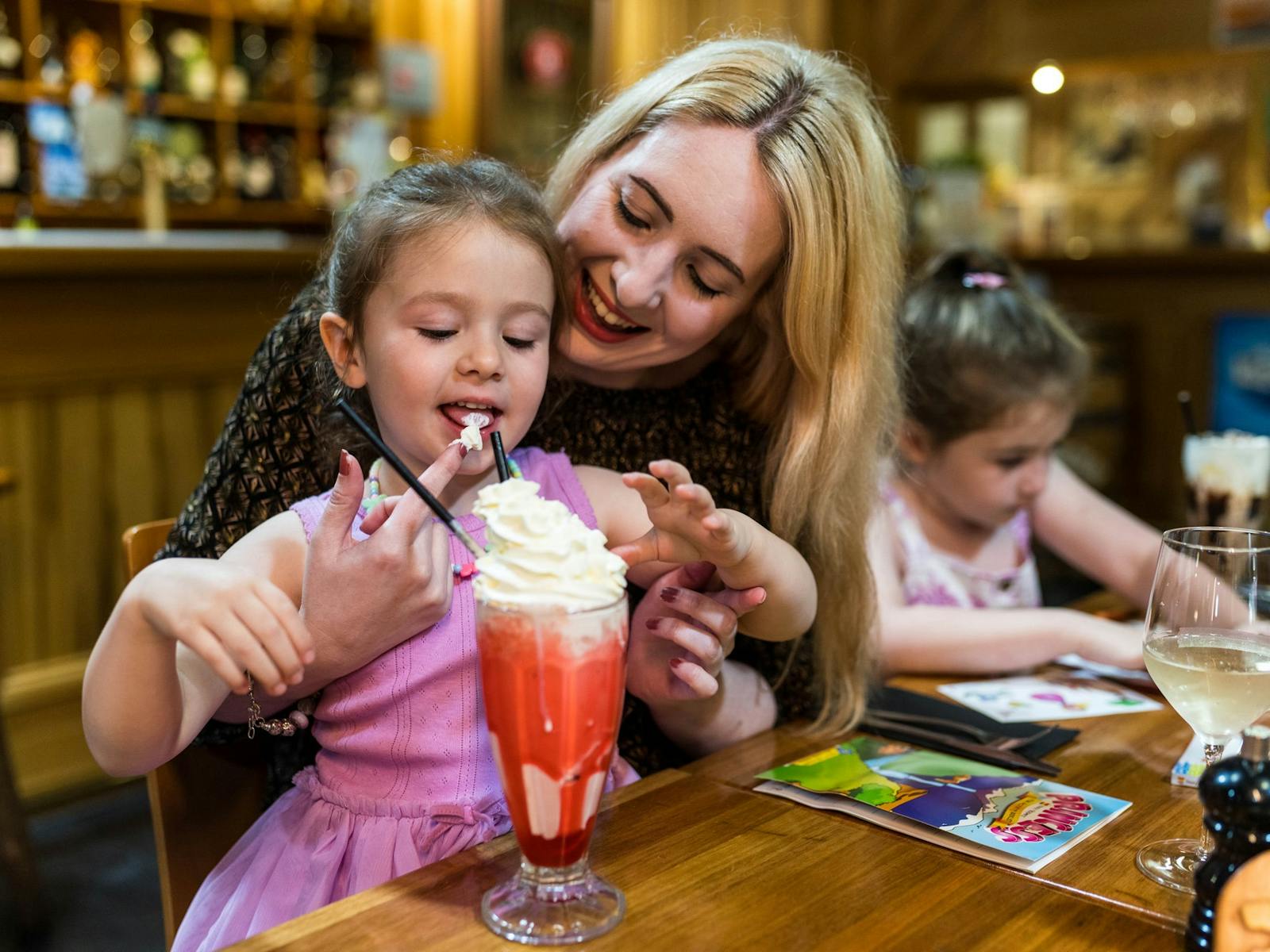 A mother smiles as she lets her youngest daughter lick whipped cream off her finger from milkshake