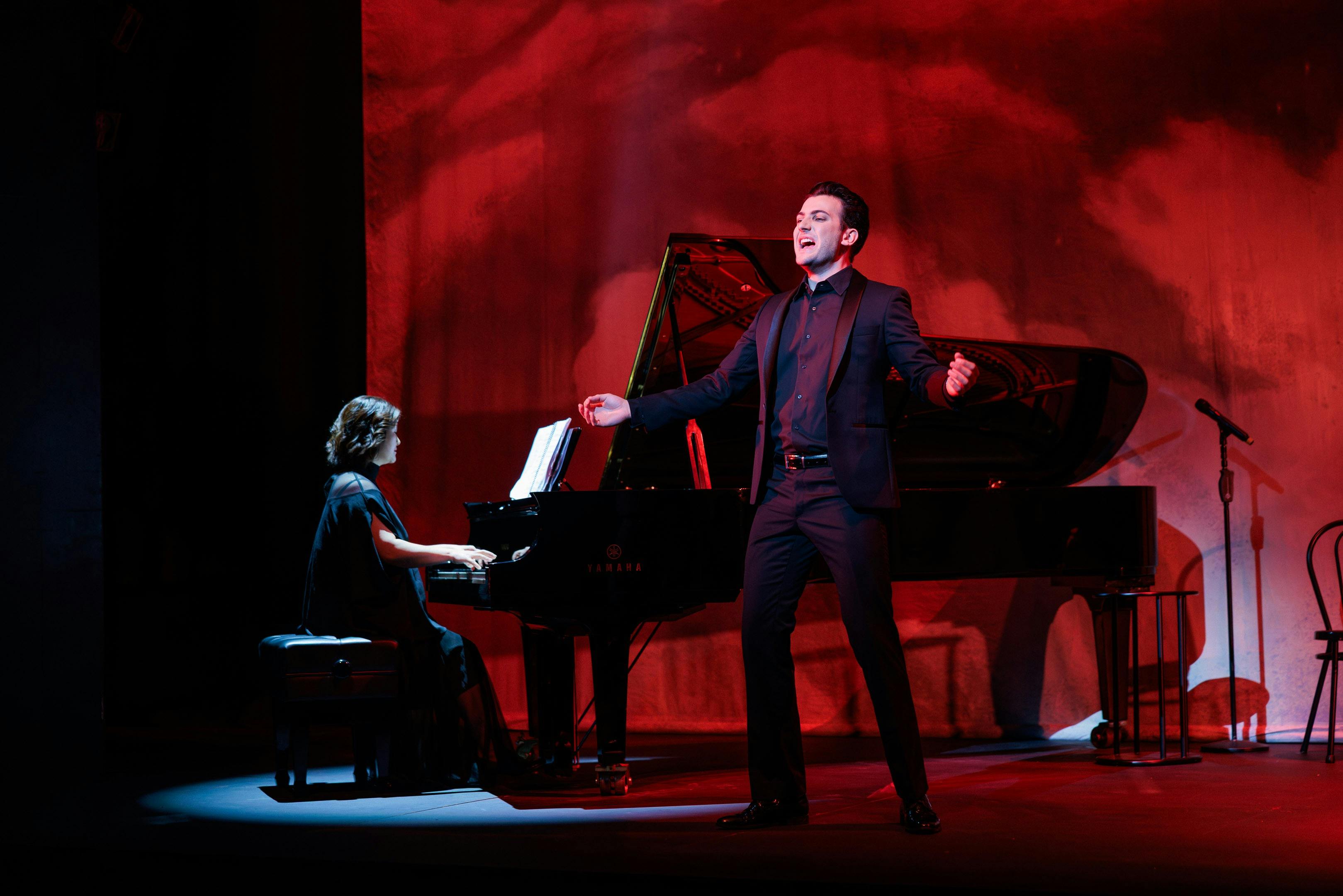 An opera singer in a black suit stands beside a piano singing