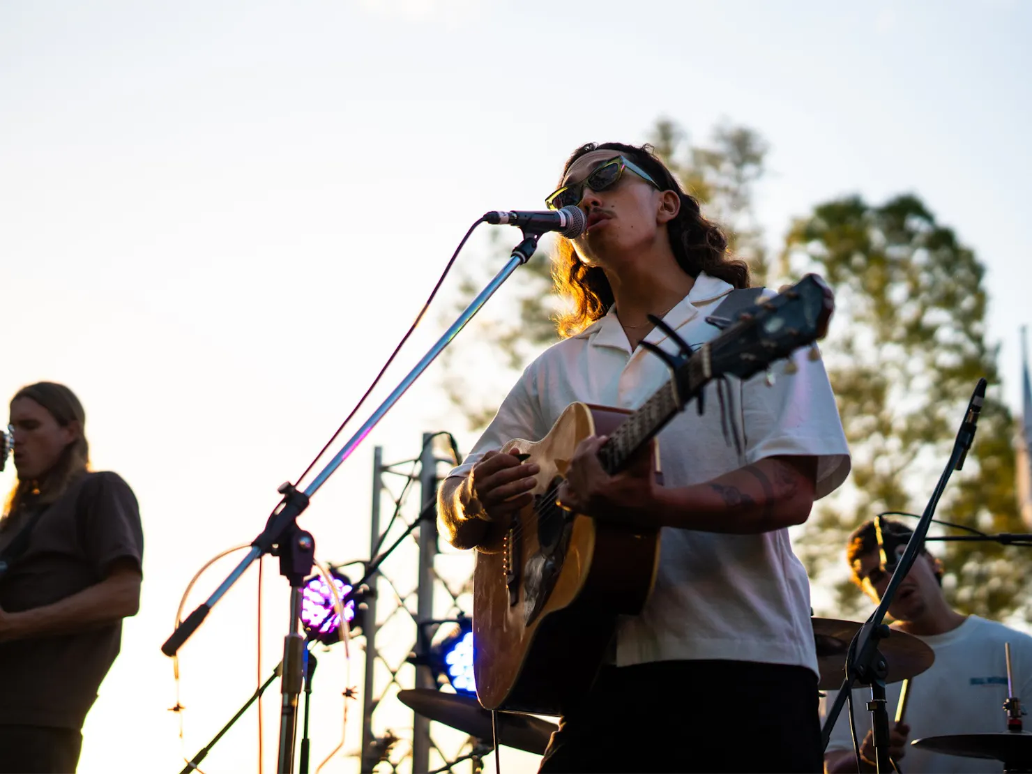 Photo of musician playing guitar and singing on stage, with another musician, tress in background
