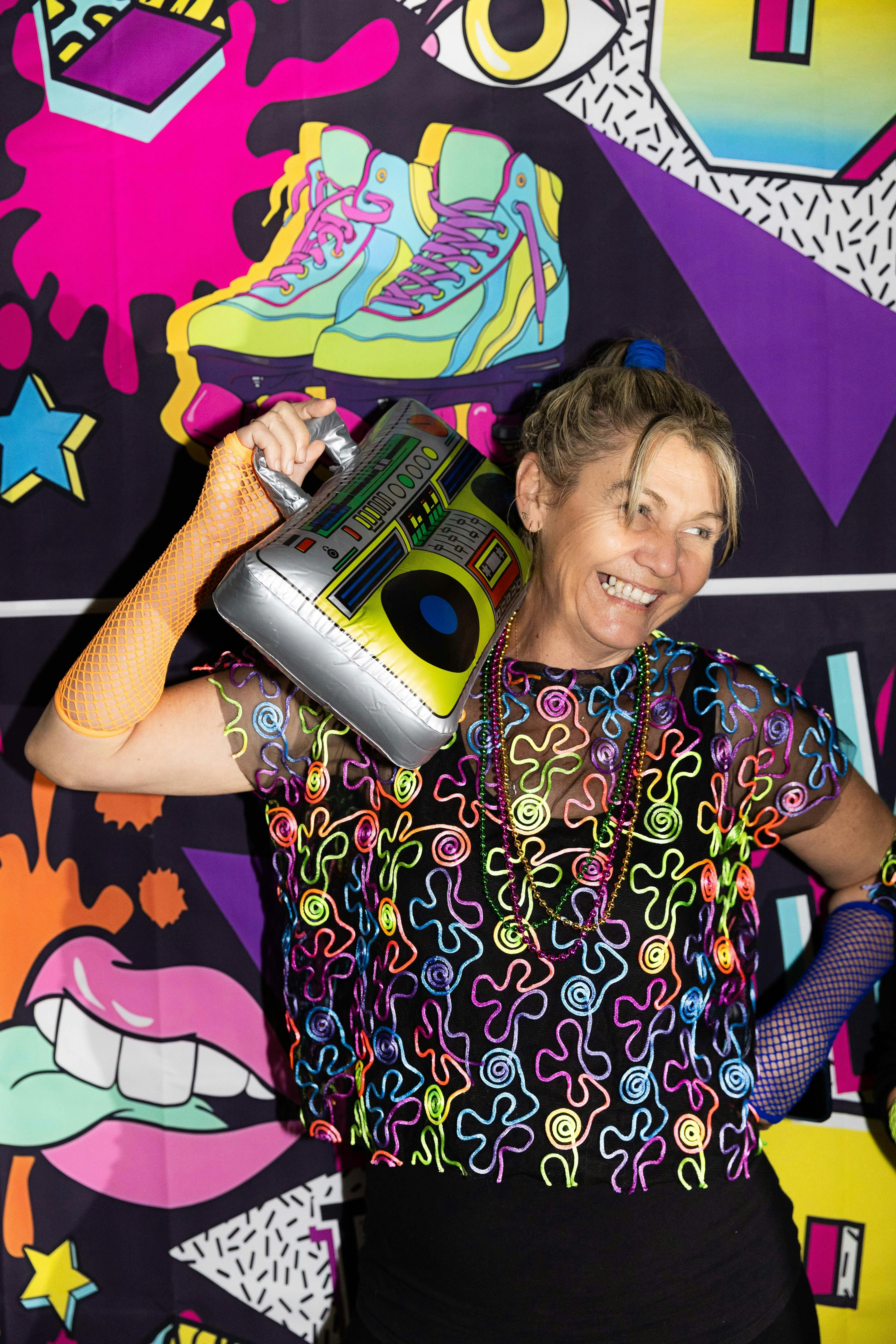 A woman in 80s costume holds a blow up cassette player against a colourful backdrop