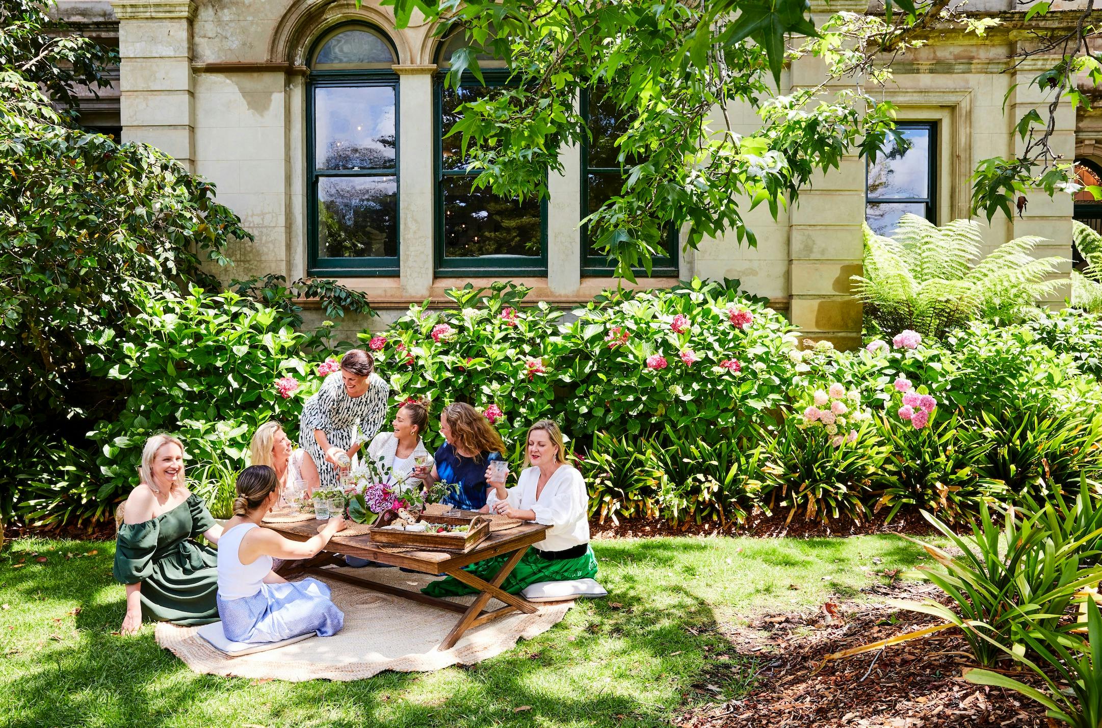 A group of women sitting on cushions at a raised picnic table  enjoying a grazing board and wine.