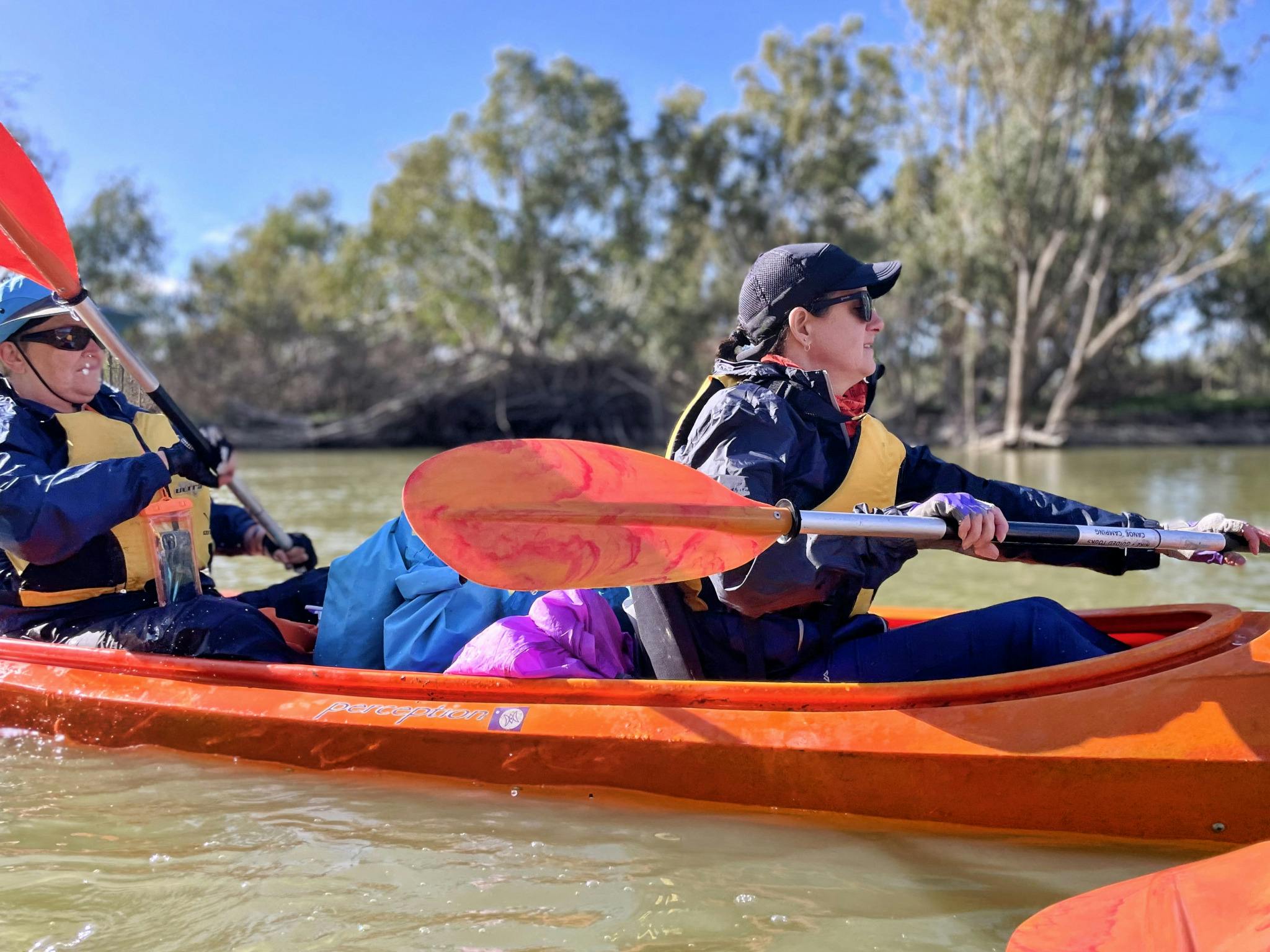 2 kayakers dressed for winter, in a loaded double kayak, paddling under a sunny sky with redgums