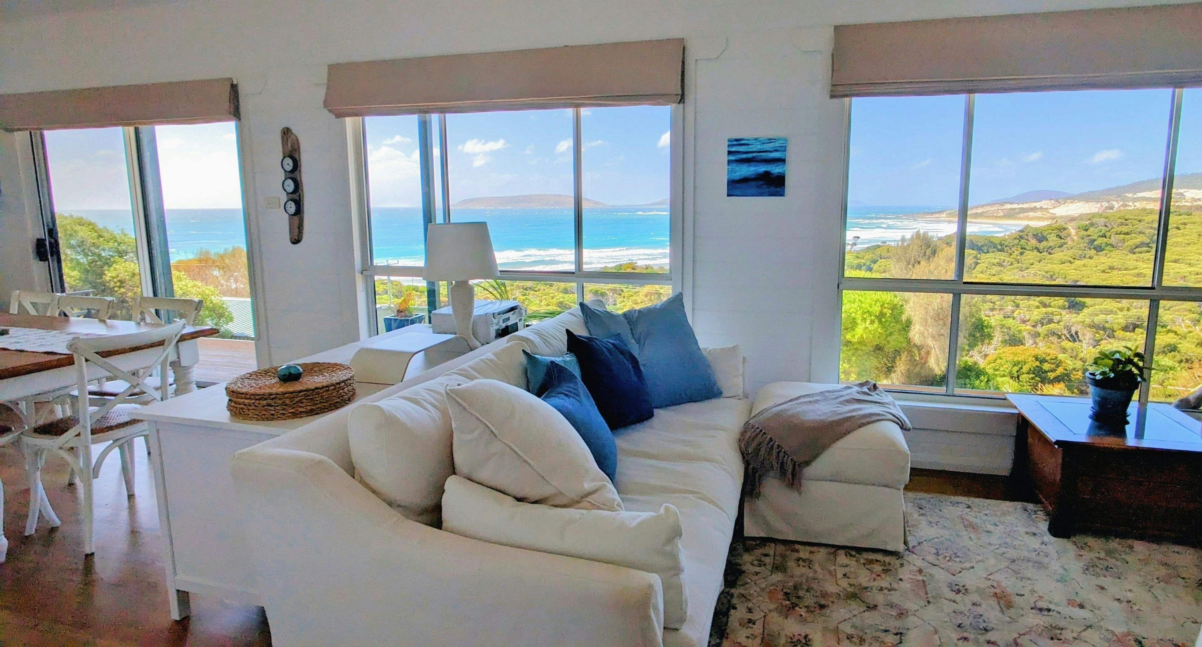 White sofa with blue cushions, lamp and large windows look out to ocean view.