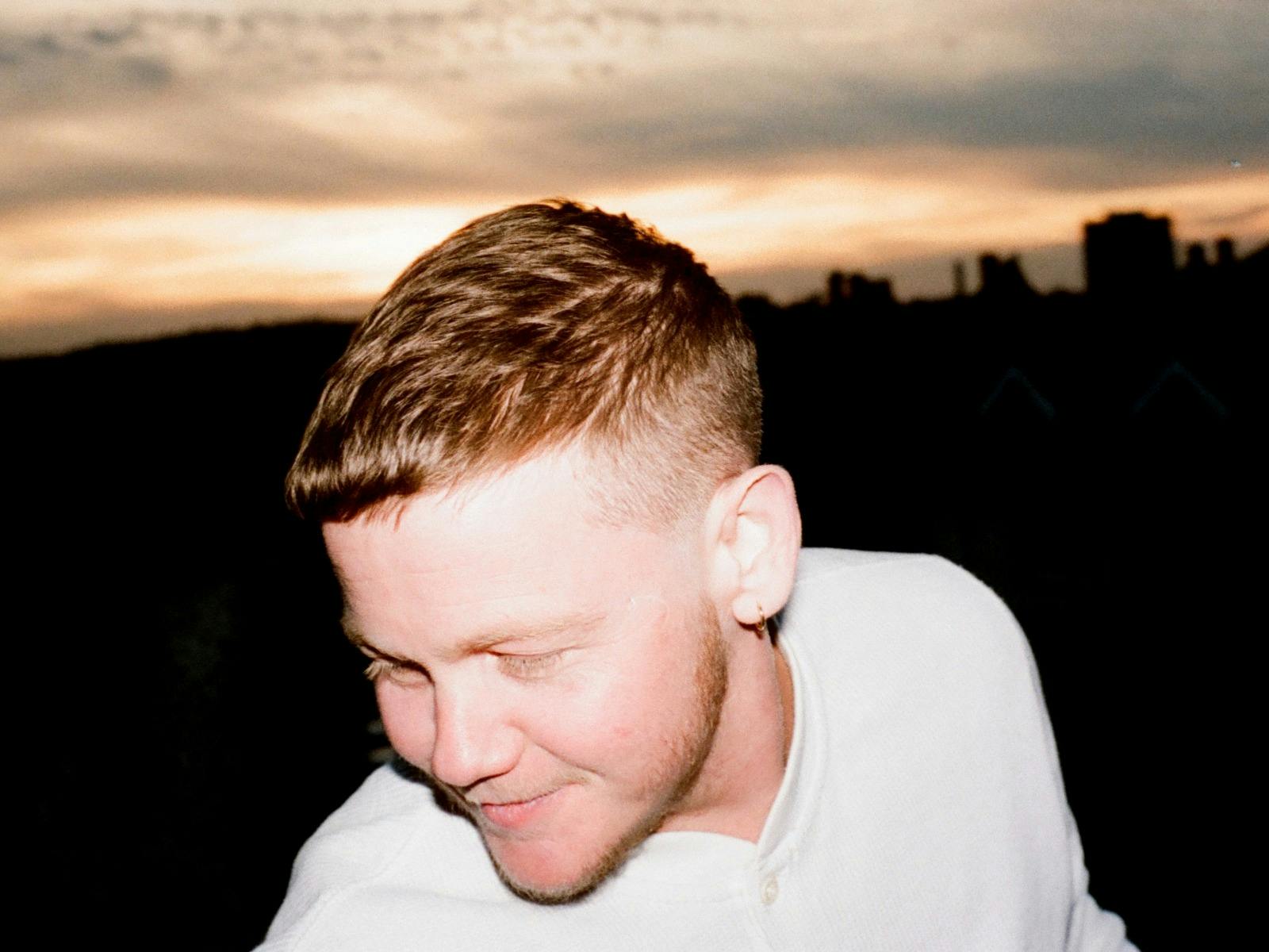 Man with short hair and earring looks down at sunset, city skyline silhouetted in the background.