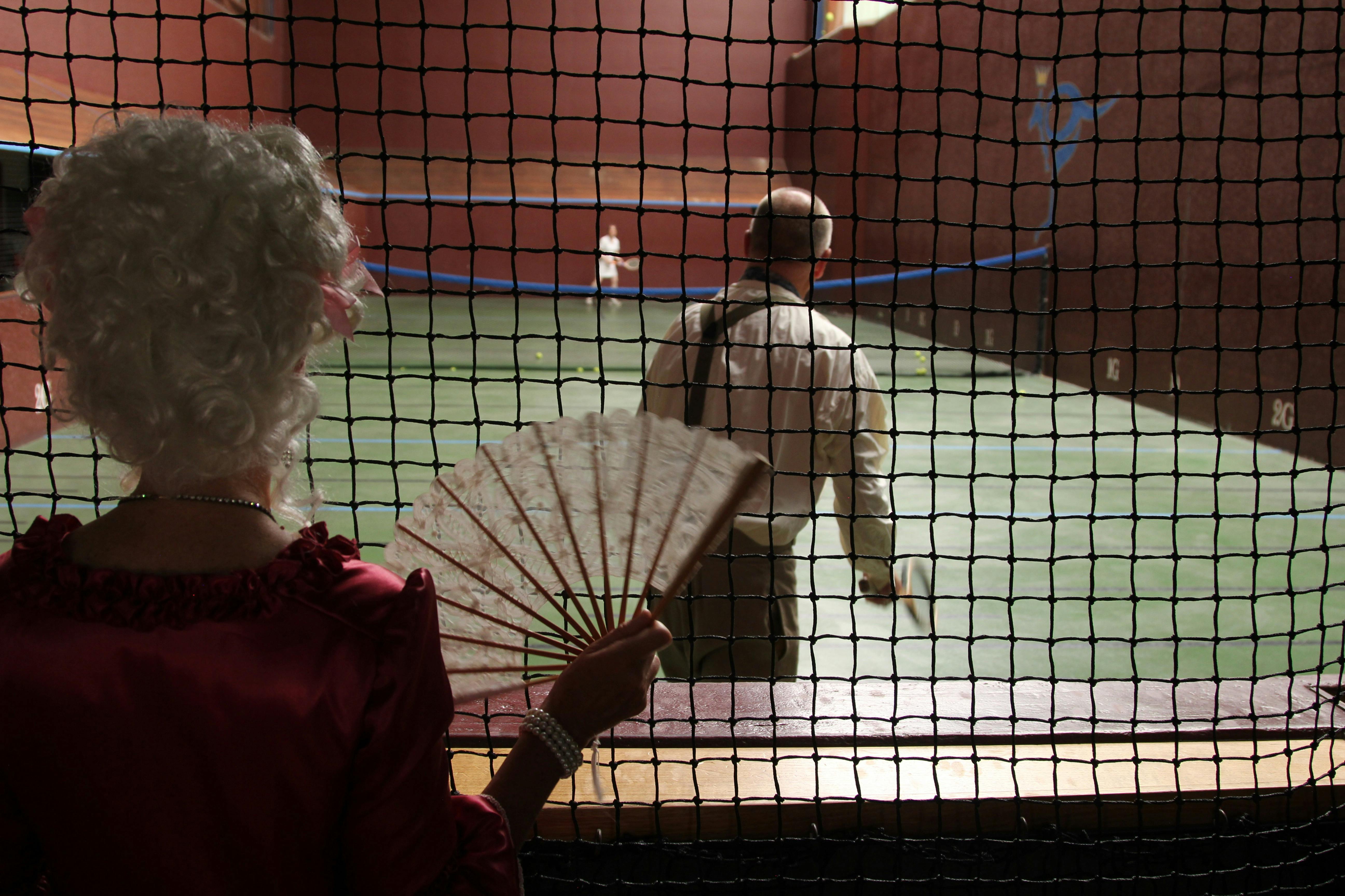 A woman in 18th century dress is waving a fan and watching a game of real tennis from the gallery.