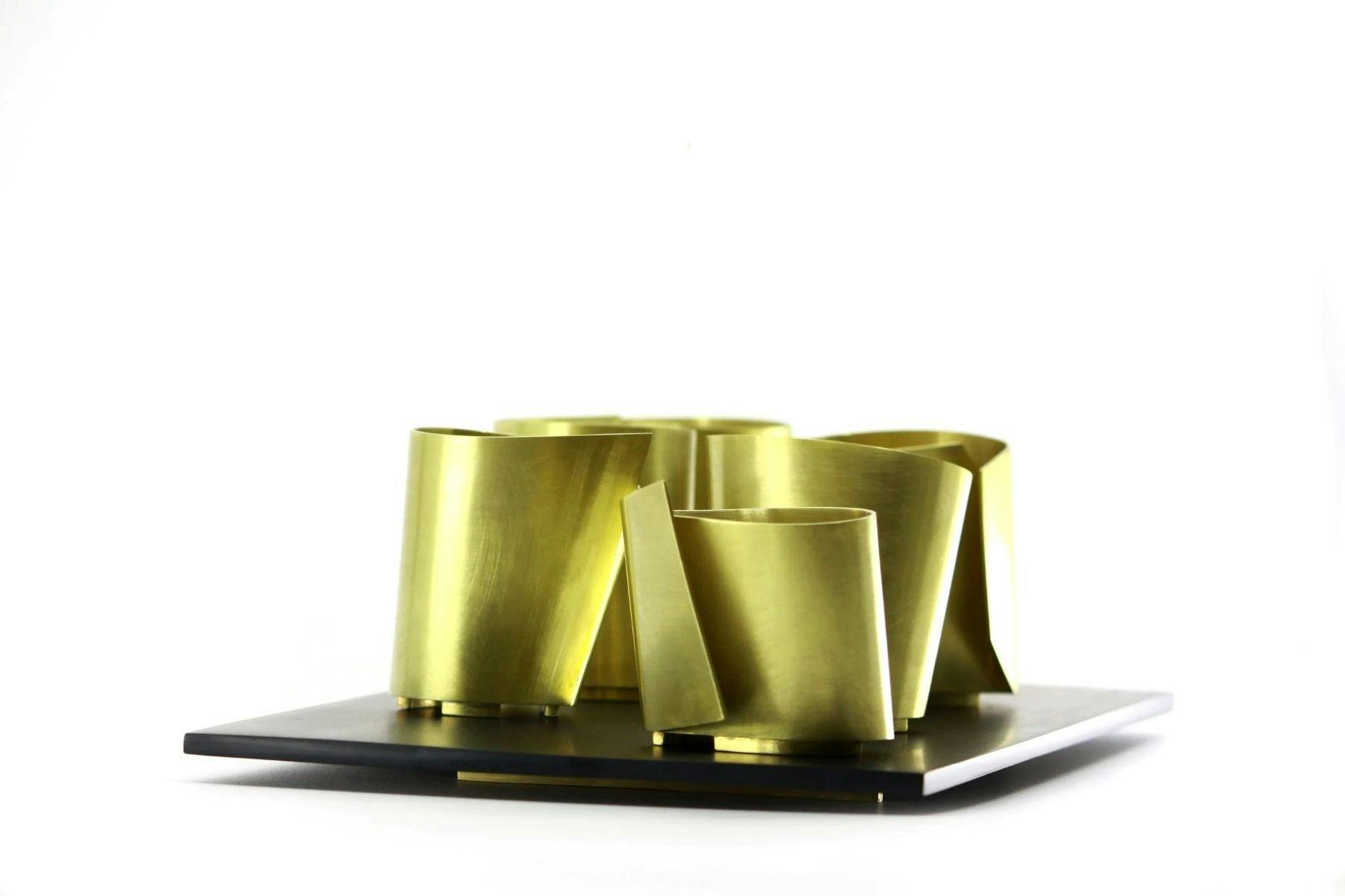 A collection of brass metal vessels with sharp angles sits on a platter against a white background