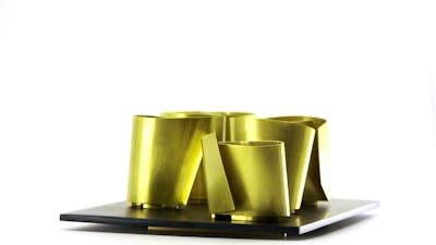 A collection of brass metal vessels with sharp angles sits on a platter against a white background