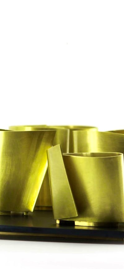 A collection of brass metal vessels with sharp angles sits on a platter against a white background