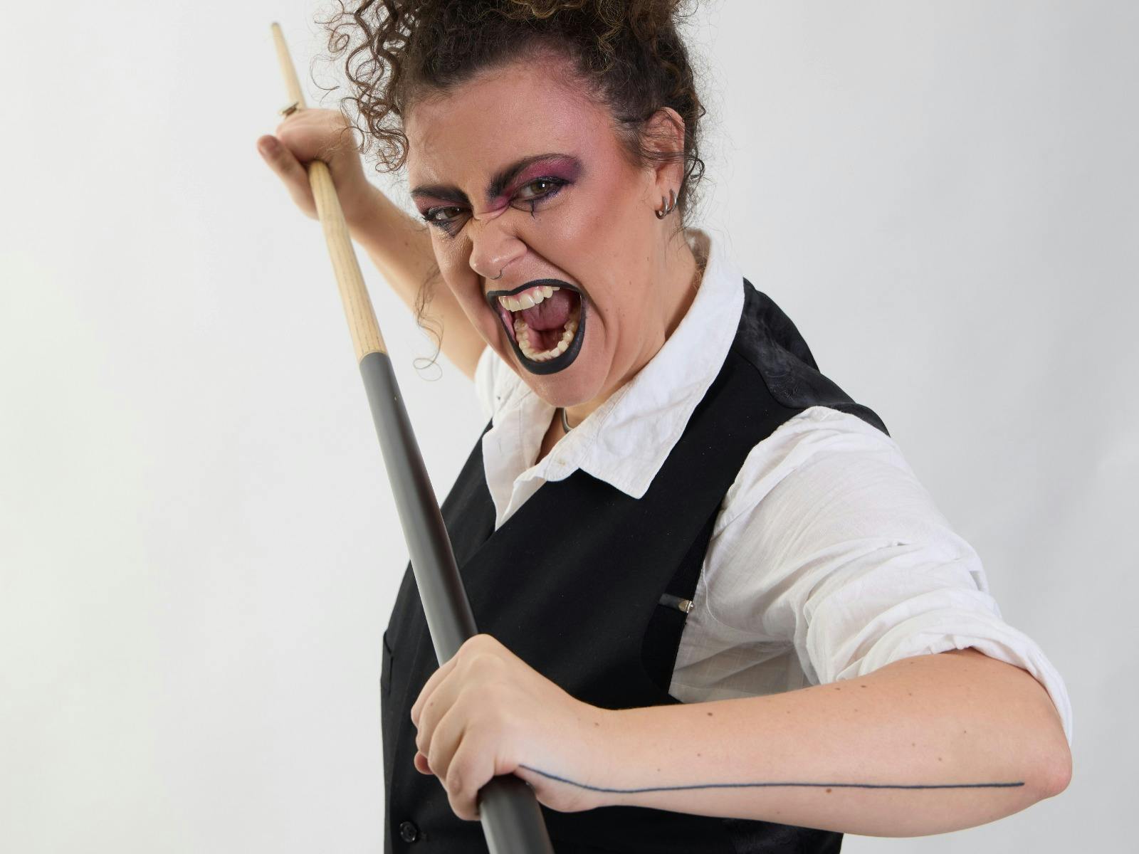 A person wearing a white shirt and black vest holds a poker cue and screams at the camera