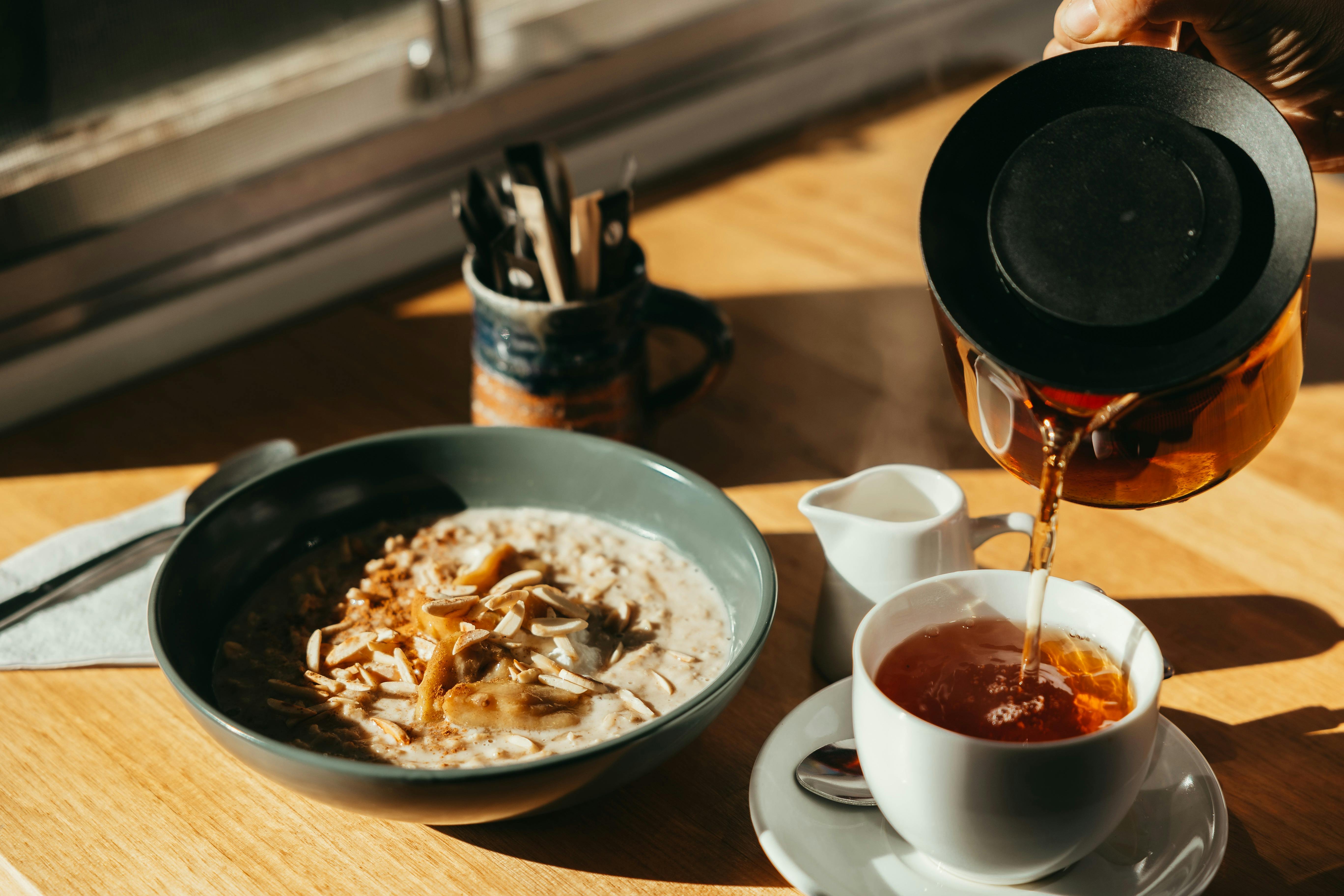 A bowl of porridge sits on a table as a cup of tea is being poured from a jug.