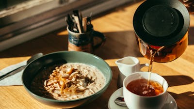 A bowl of porridge sits on a table as a cup of tea is being poured from a jug.