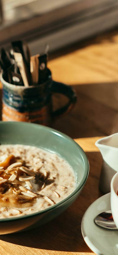 A bowl of porridge sits on a table as a cup of tea is being poured from a jug.