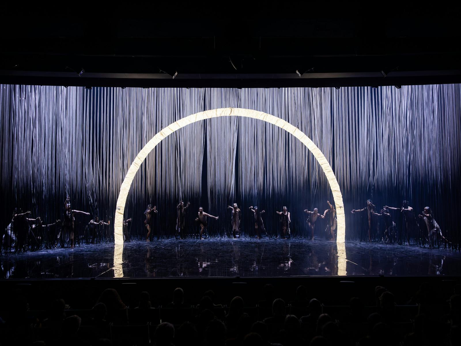 A troupe of dancers stand behind a roped curtain and reach through. There are is a large light arch.
