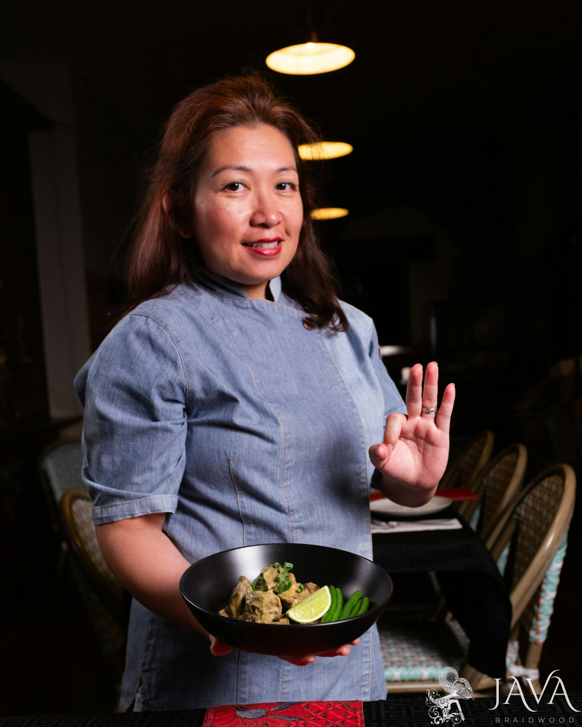 Chef Daniar warm evening lights, denim chef attire, holding gule domba (lamb gule) in a black bowl..