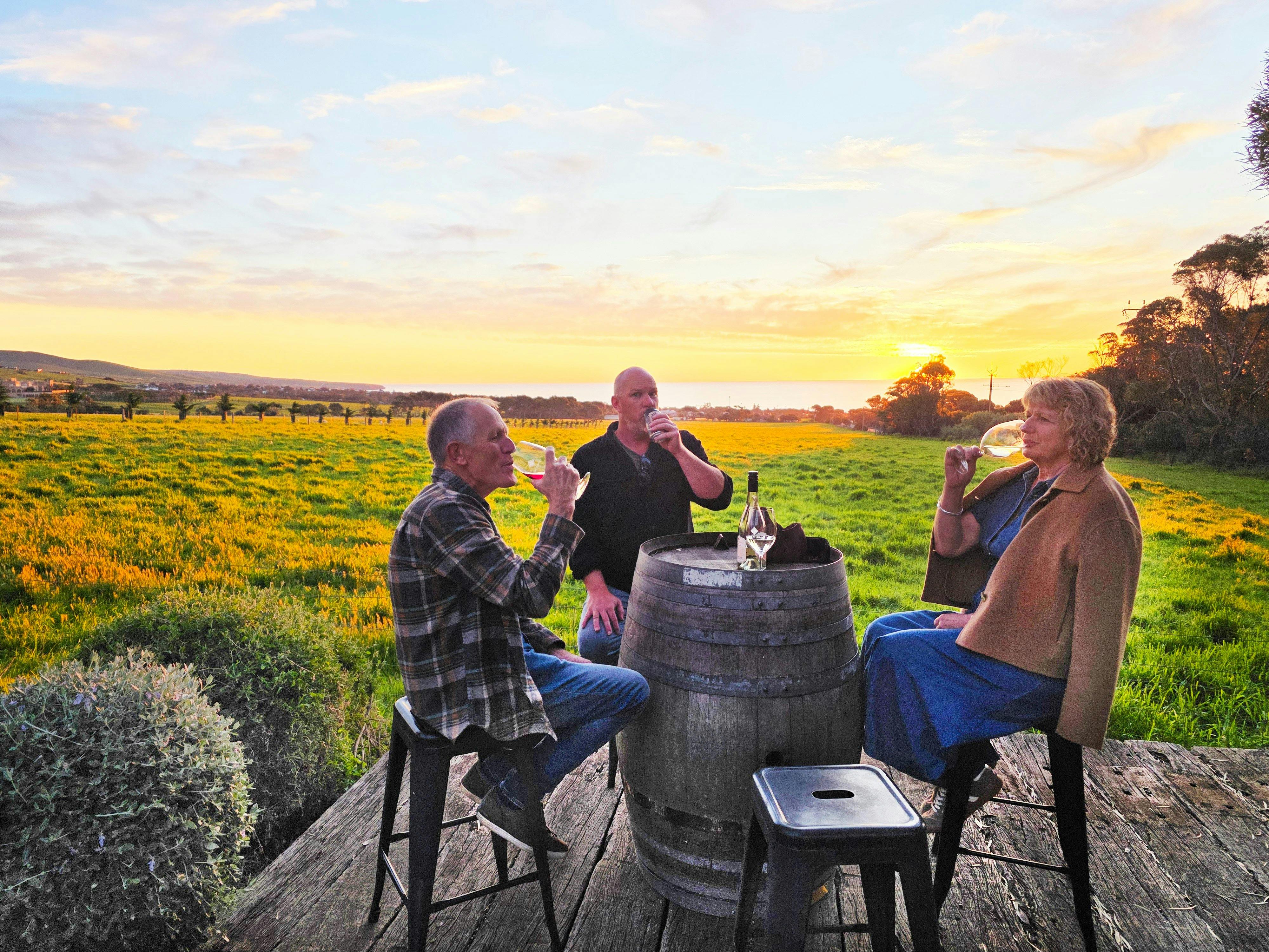 3 people sitting at a wine barrel drinking wine at sunset, overlooking the hills and ocean