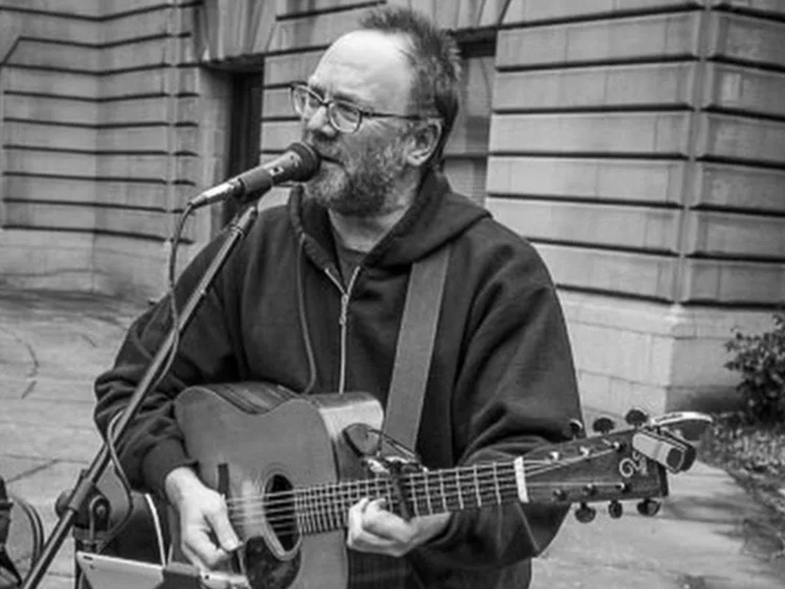David Rovics in black and white singing and playing the guitar in an outdoor space.