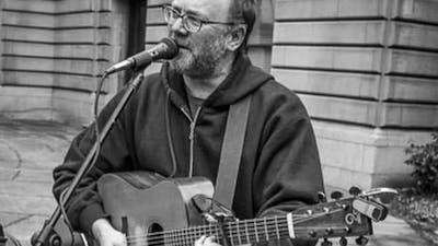 David Rovics in black and white singing and playing the guitar in an outdoor space.