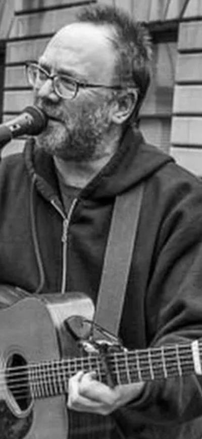 David Rovics in black and white singing and playing the guitar in an outdoor space.
