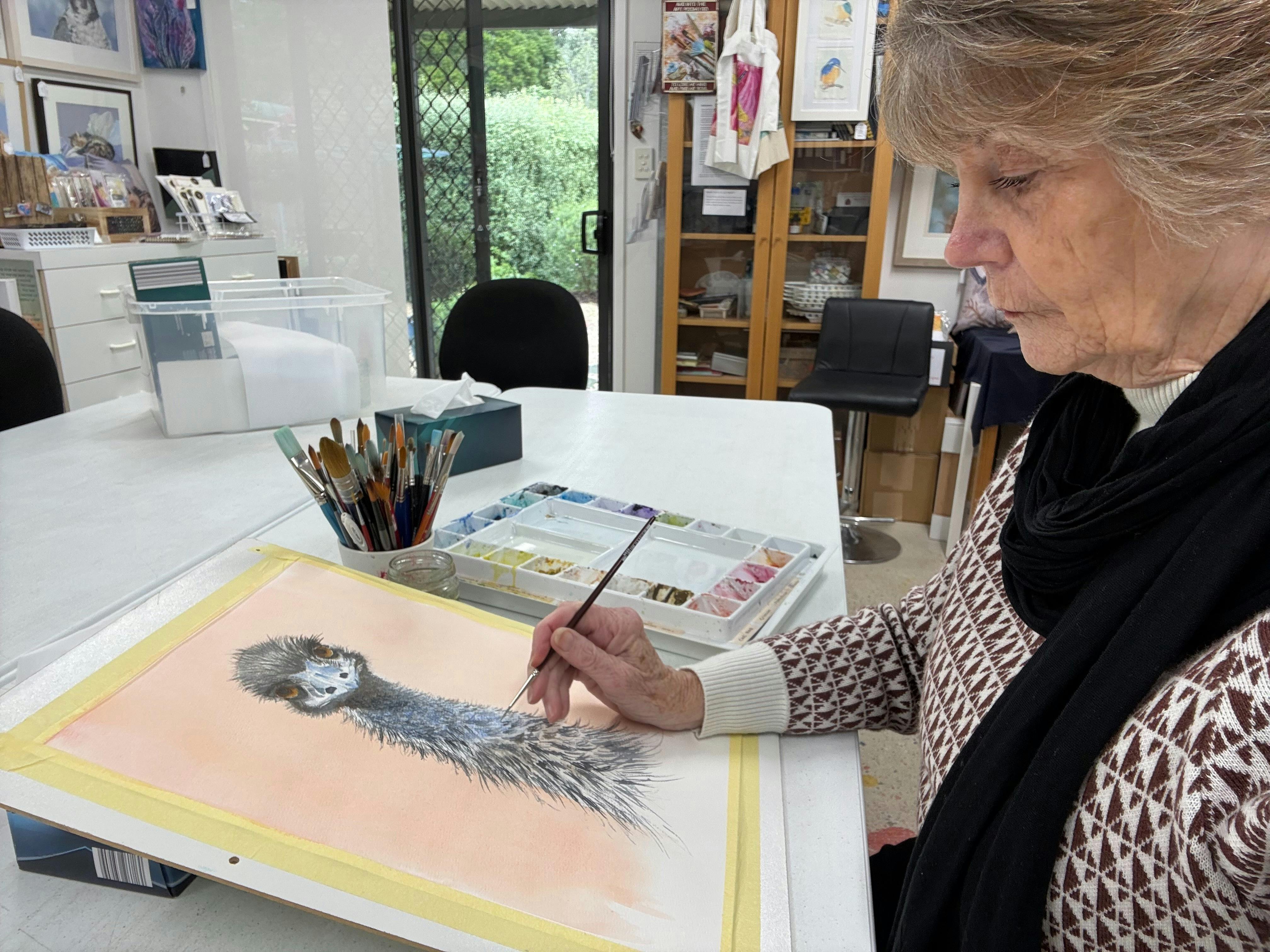 Artist in her studio painting a detailed watercolour of an emu's head and neck feathers.