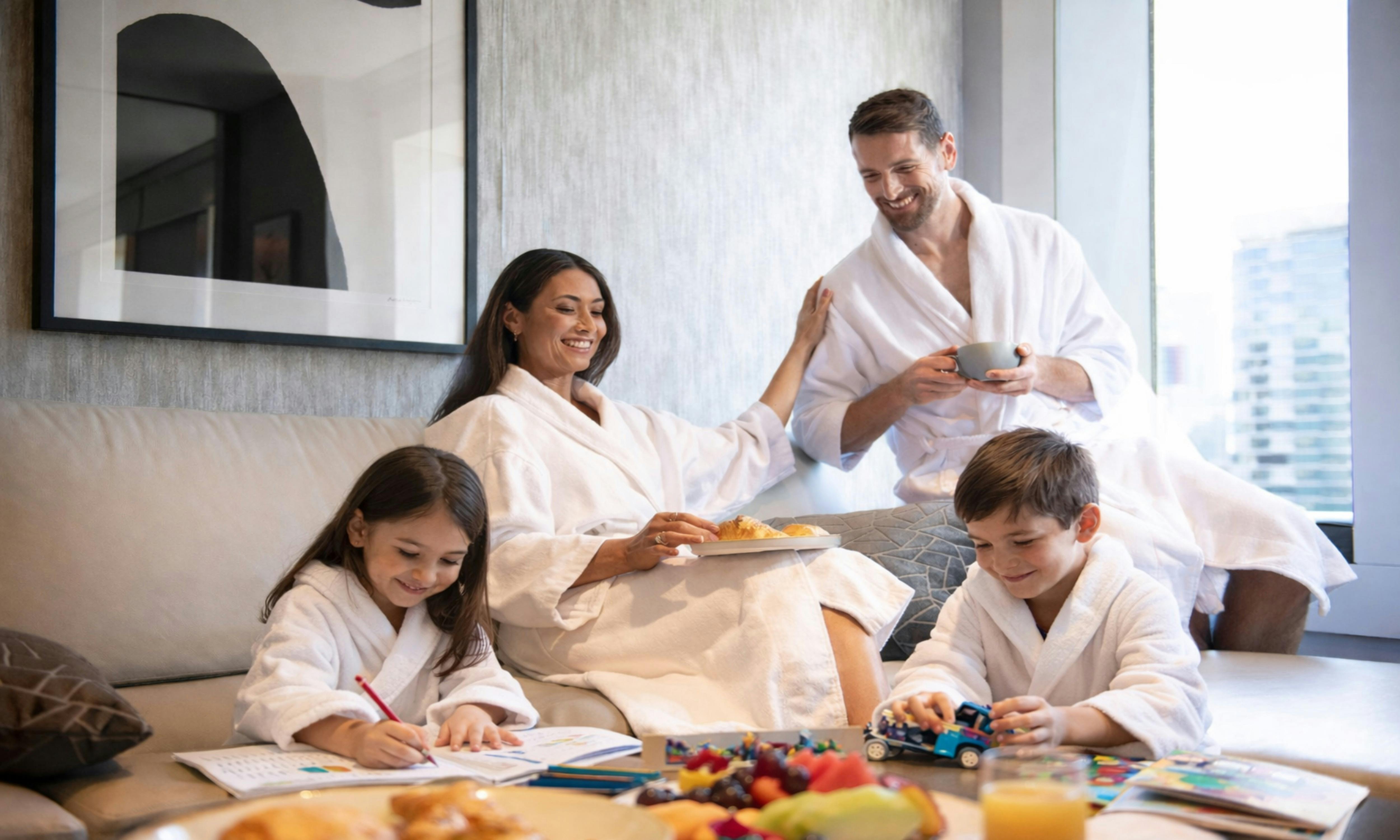 Two parents enjoy breakfast in their room as their two young kids colour in and play in the room.