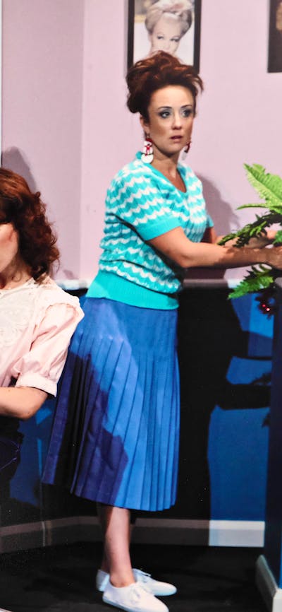 three women in a 1950s hair salon, one has her hair in curlers under the dryer