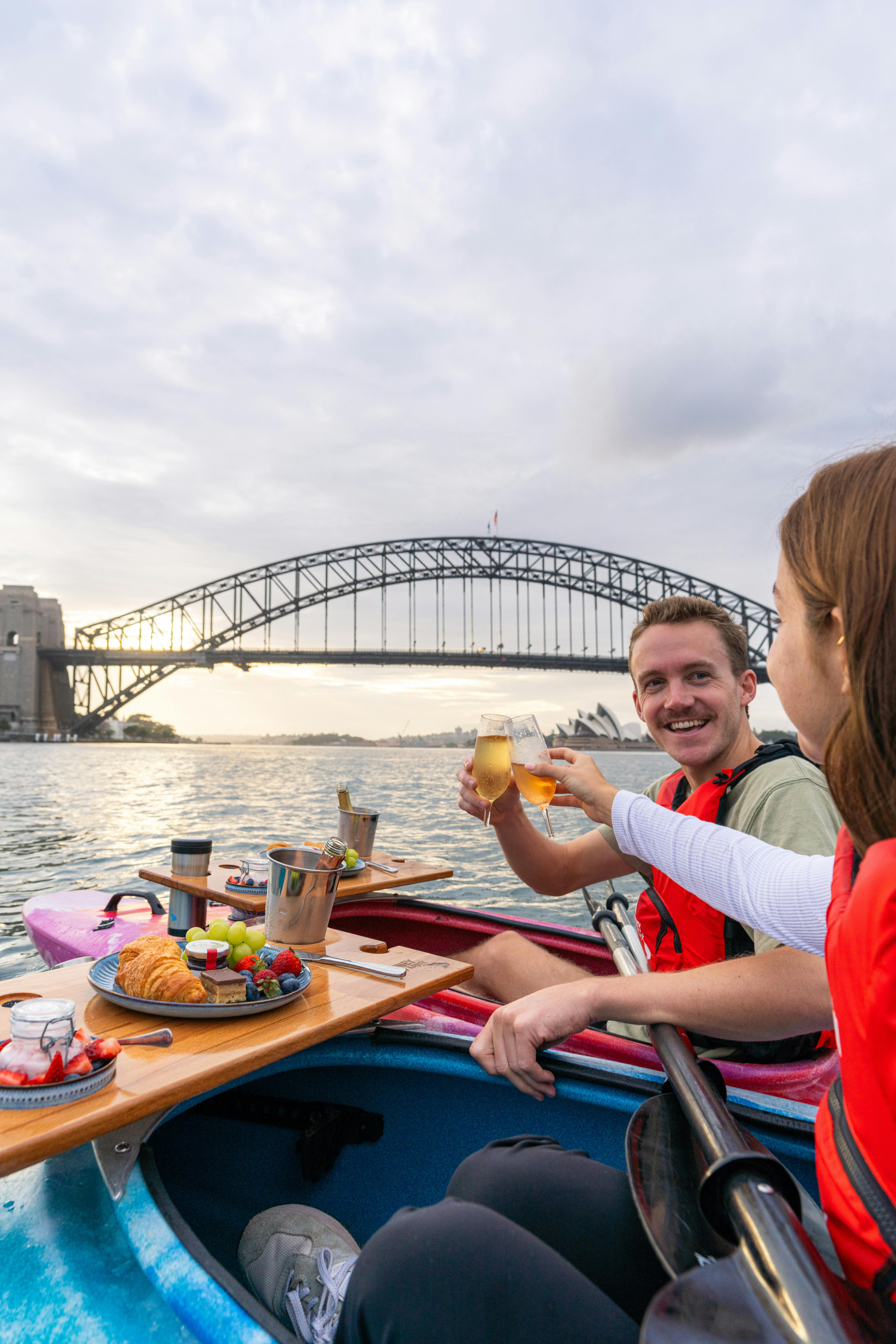 Couple enjoy champagne breakfast from their kayaks viewing Sydney harbour bridge and the opera house