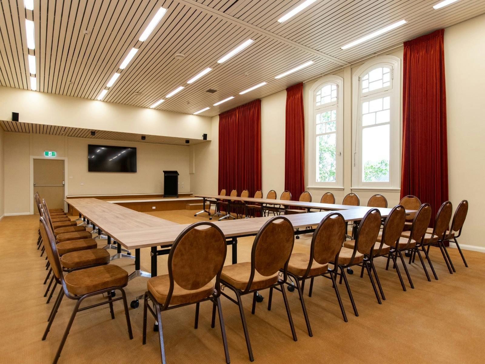 A carpeted room with chairs and tables, a screen and floor to ceiling curtains
