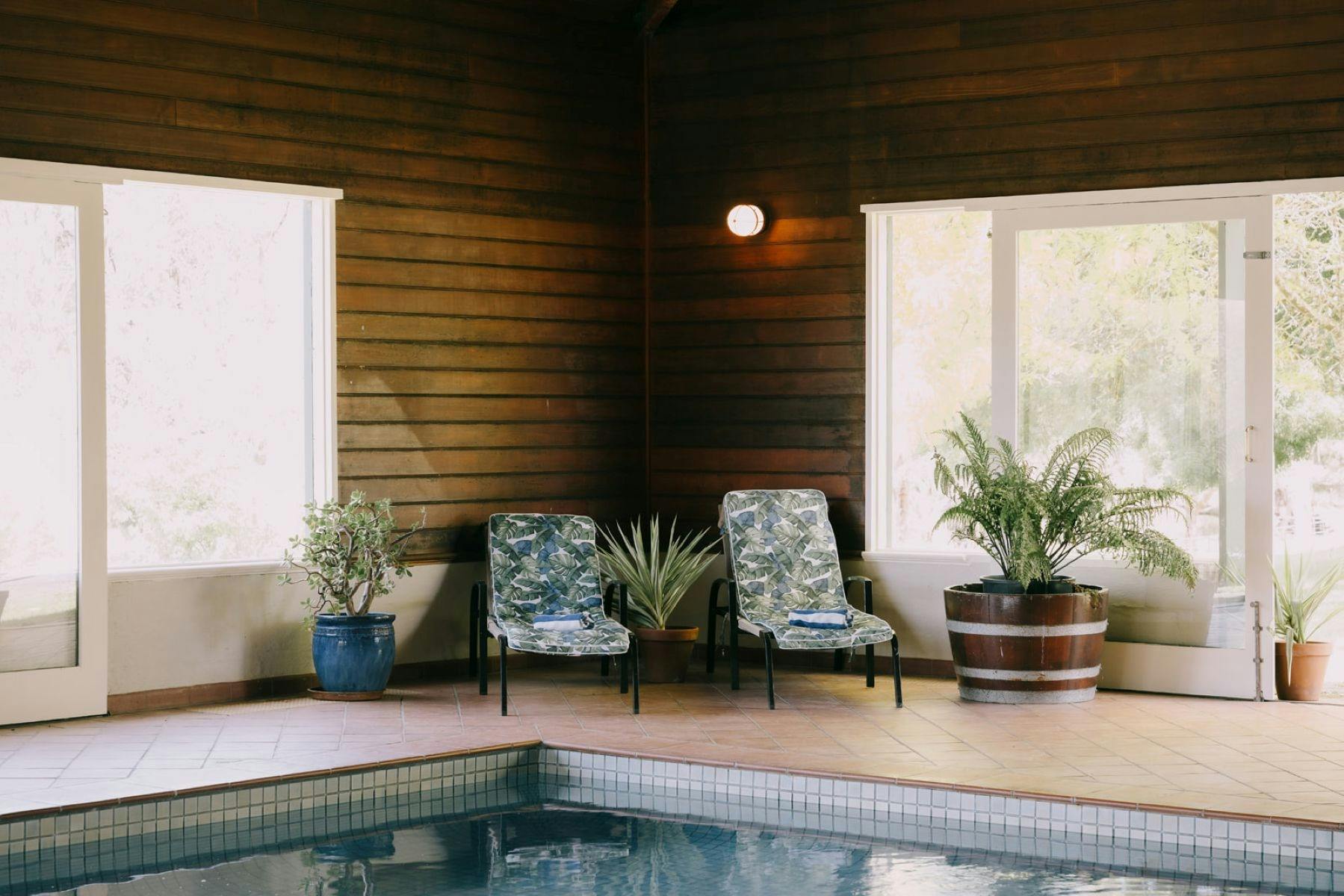 Indoor pool with lounge chairs, potted plants, wooden walls, and large windows letting in natural li