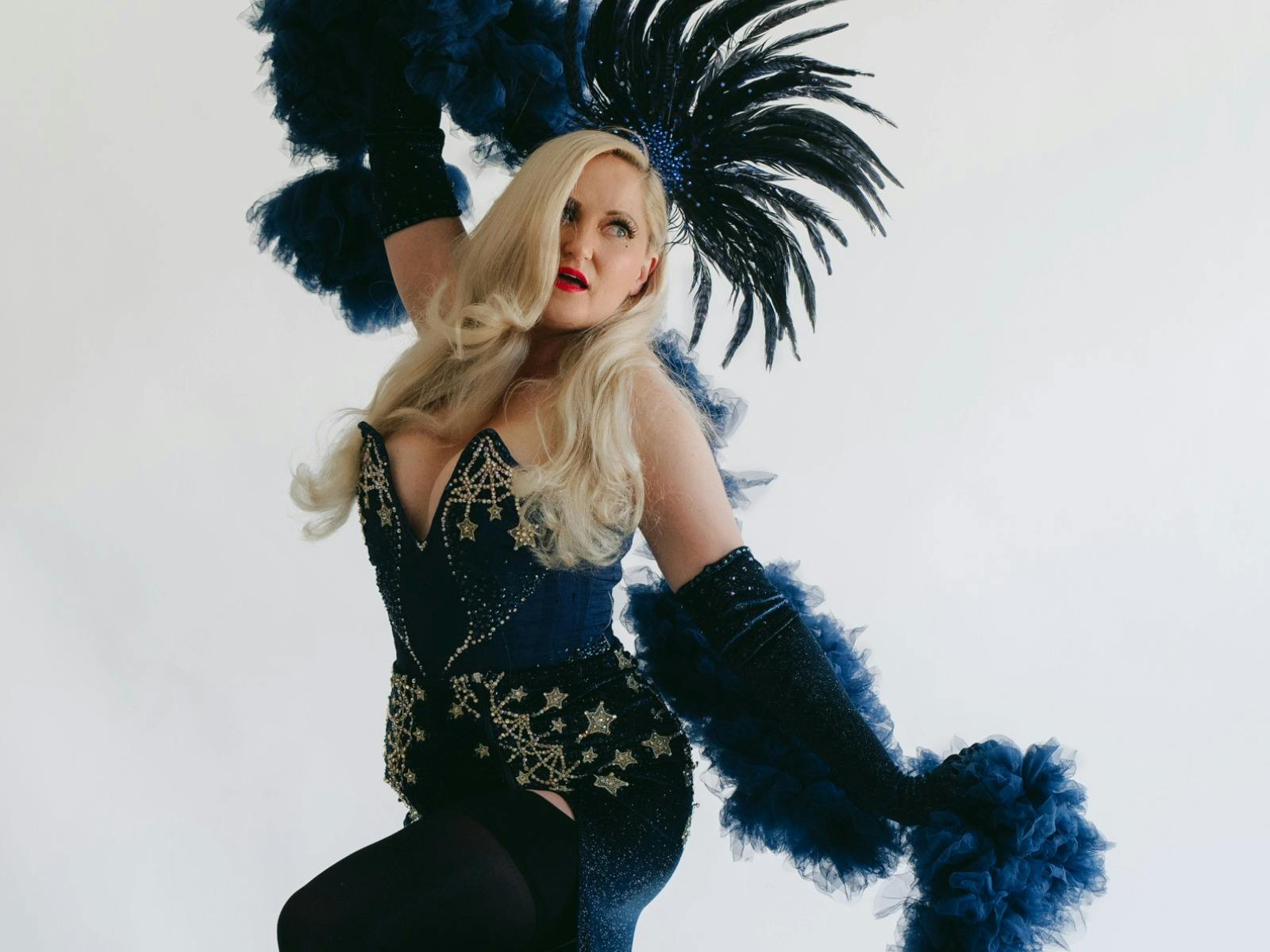 Lyra La Belle is holding her blue boa with one arm above her head, the other out to her side
