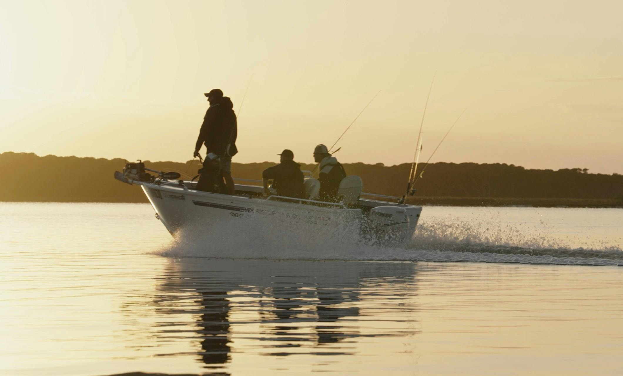 Three anglers in a boat silhouetted against a golden sunrise on calm water