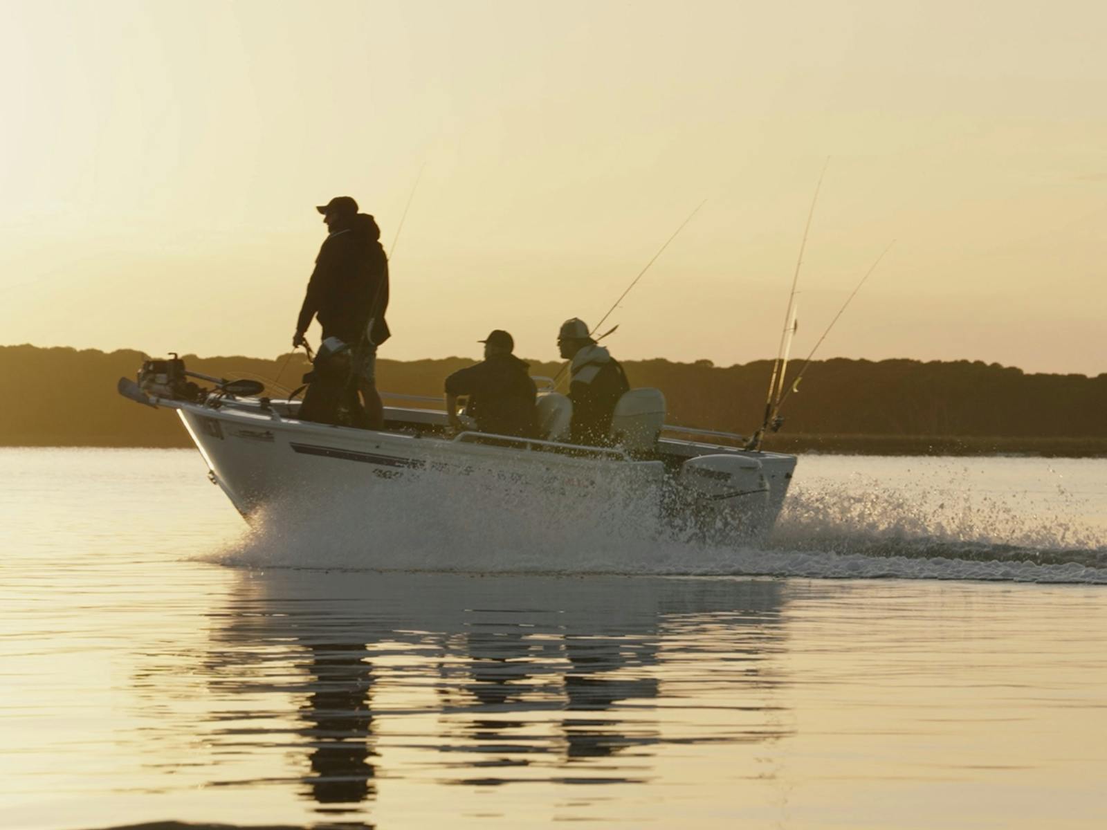 Three anglers in a boat silhouetted against a golden sunrise on calm water
