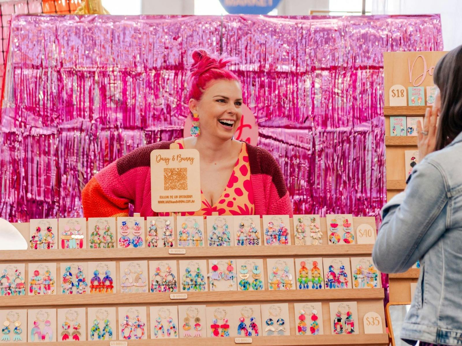 A woman with pink hair stands behund her stall laughing with someone off camera