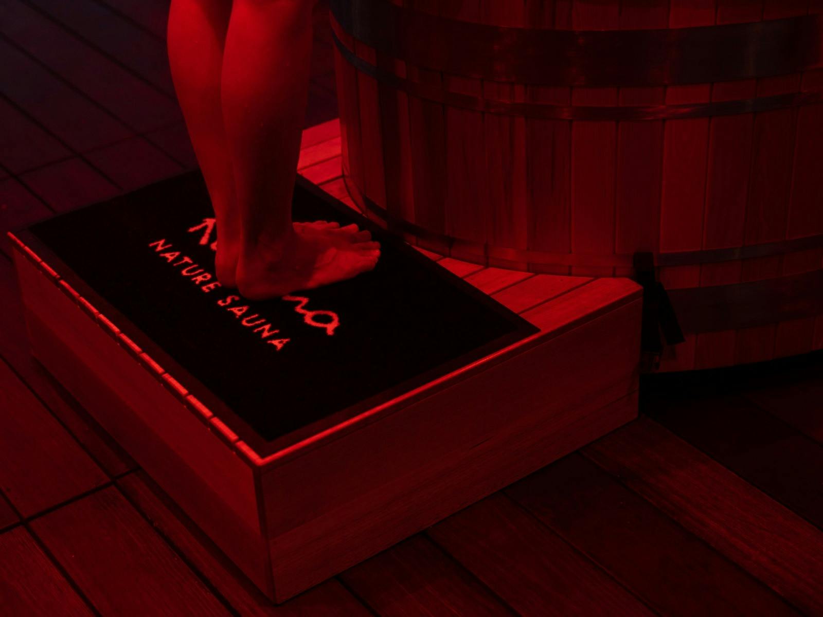 Bare feet on a "KUUMA" mat next to a wooden hot tub in deep red light.