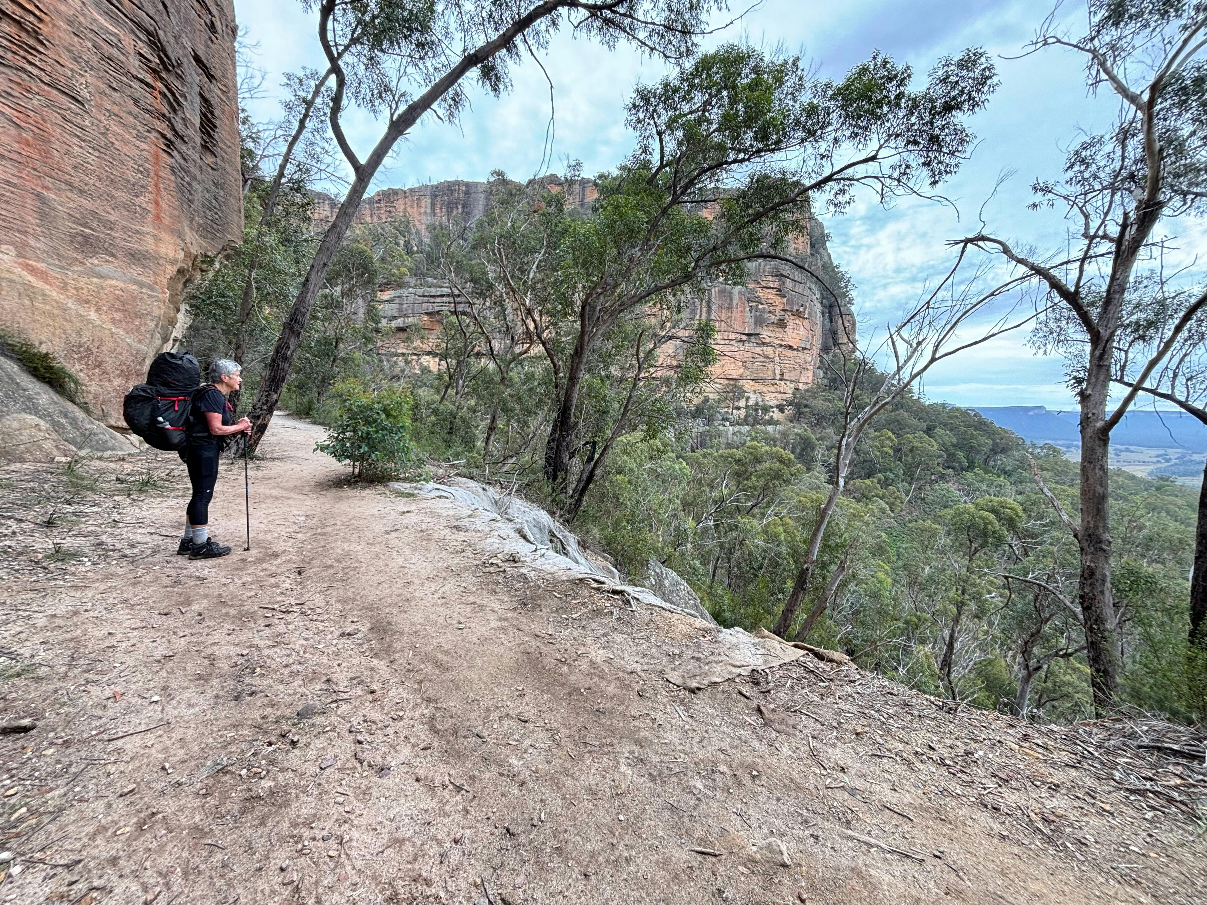A woman wearing a back pack standing beneath a sand stone cliff and looking down into the Wolgan Val