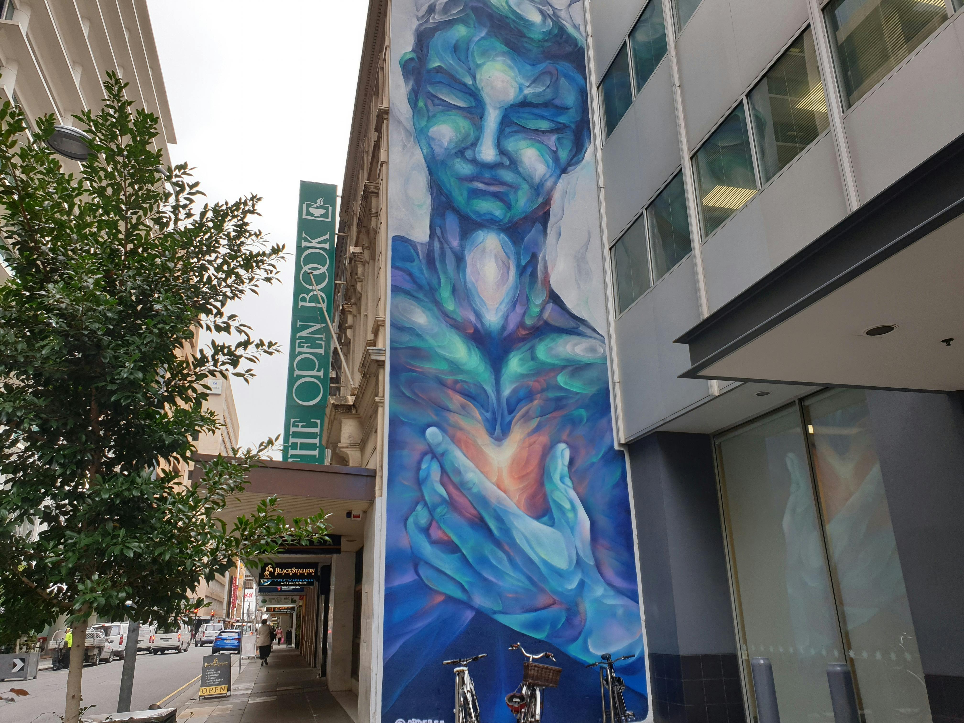 A vertical street art mural features a blue-coloured being with a hand holding its heart.