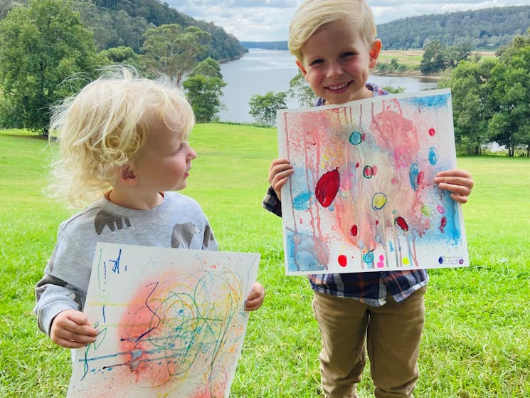 Two children stand outside holding their drawings up, behind them is a river