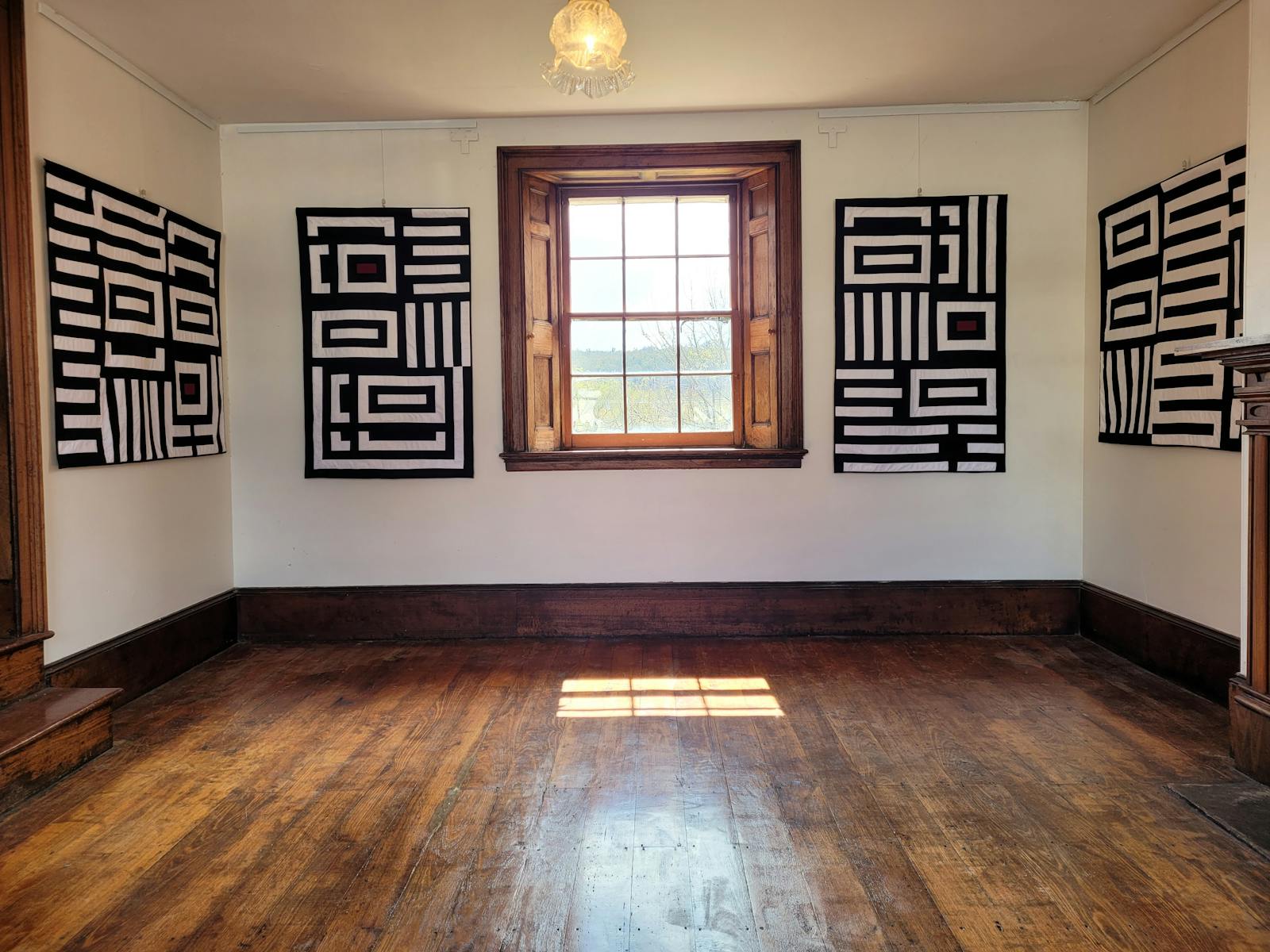 Four black and white quilts hanging in a white room with timber floor and large window.