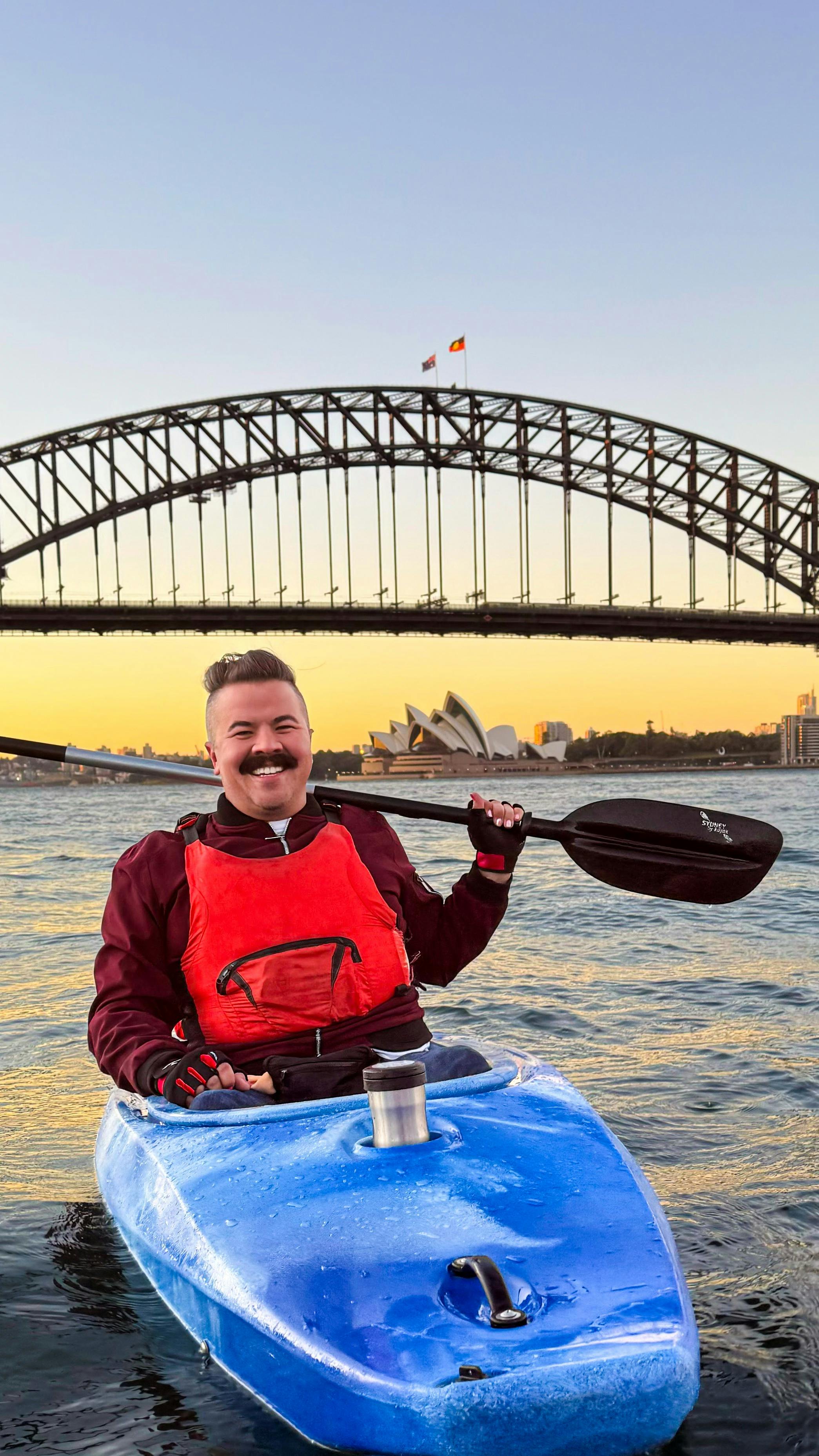 シドニー港でカヤックに座り、コーヒーを飲みながらパドルを肩に担いでいる男性