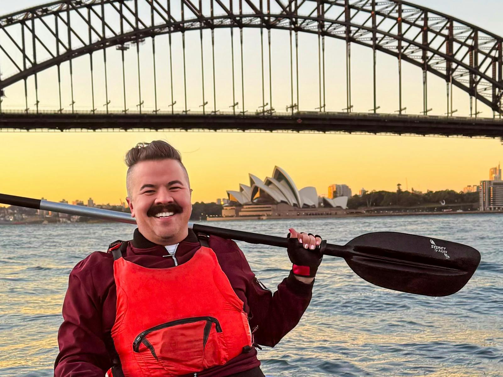 Man holding paddle on shoulder while sitting in kayak on sydney harbour with coffee in his kayak