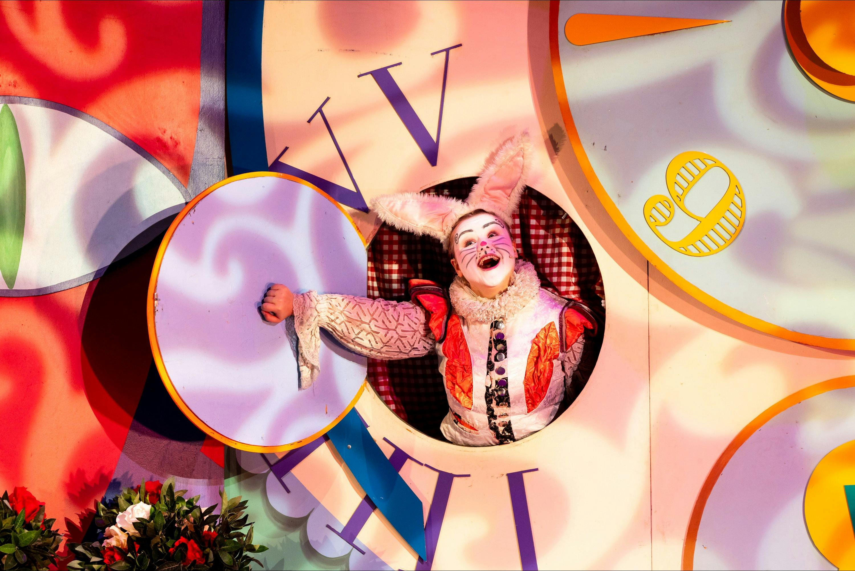 Performer dressed as the White Rabbit pops out of a clock door onstage, smiling brightly.