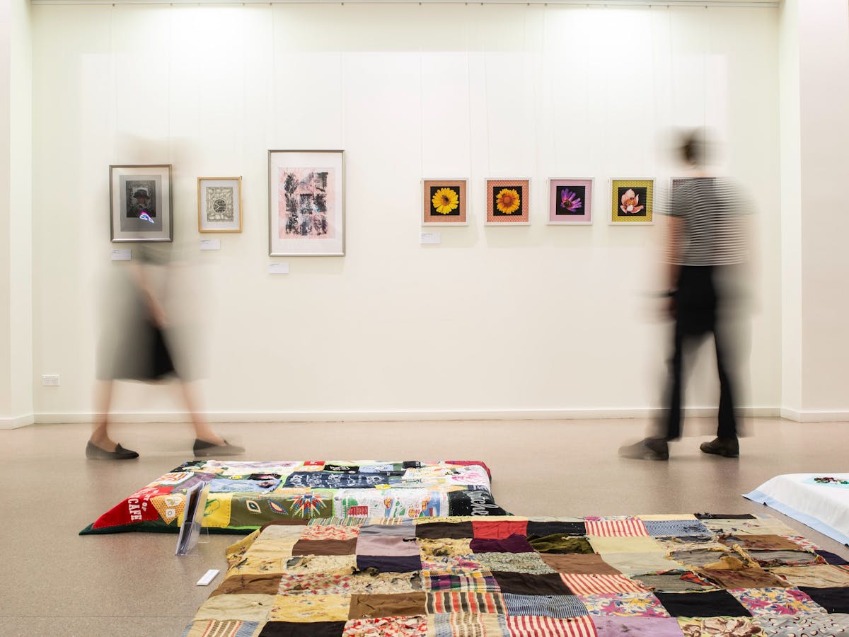 2 people are slightly blurry observing art work on the wall and on the floor.
