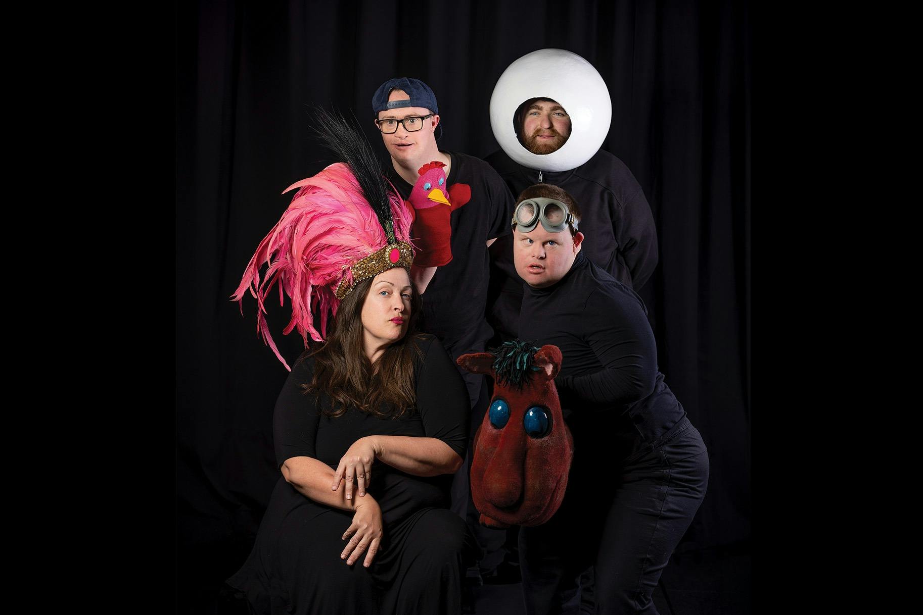 Four performers dressed in black on a black background. Each one folding or wearing a colourful prop