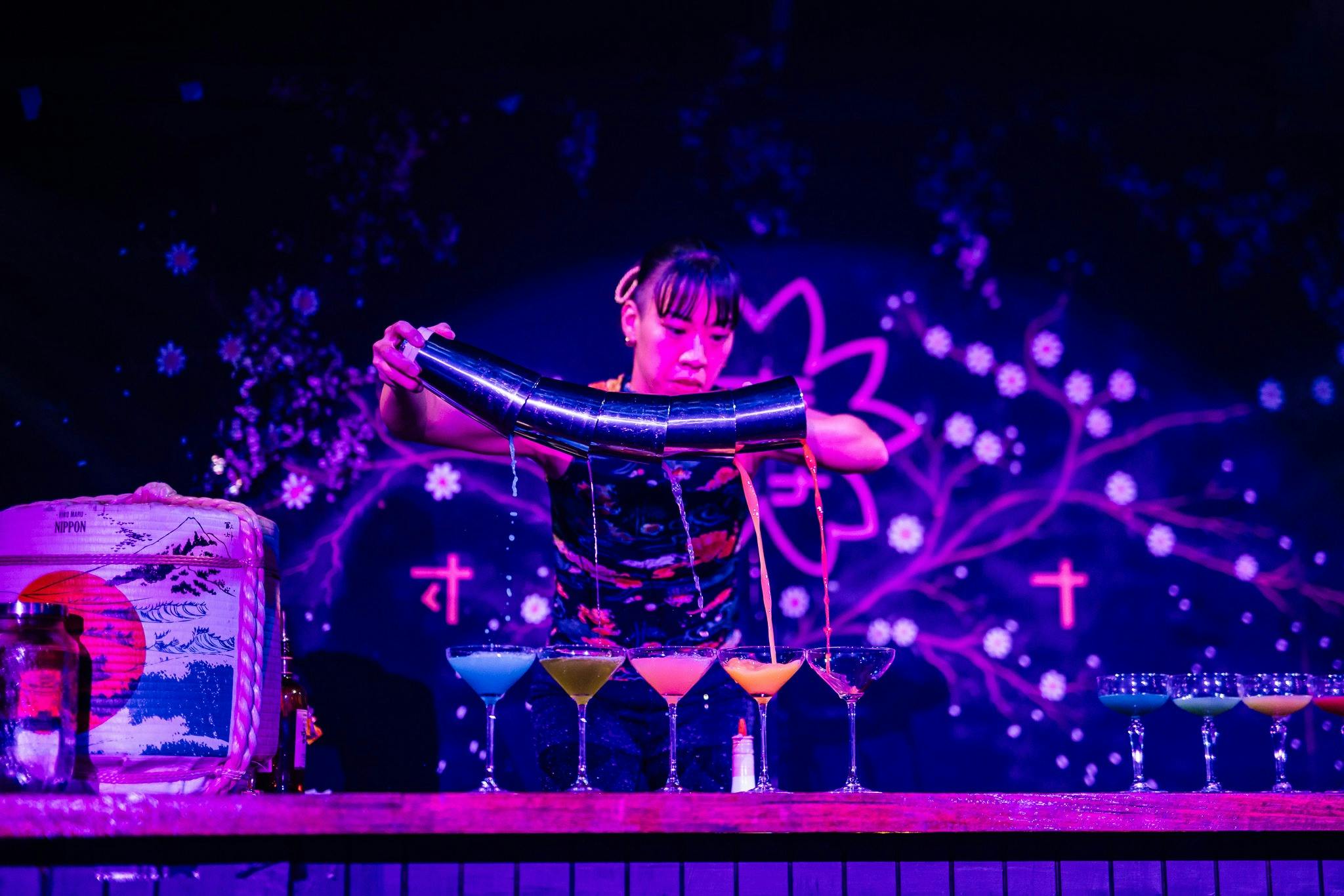 A woman pours 6 drinks at once from cocktail shakers, making a rainbow