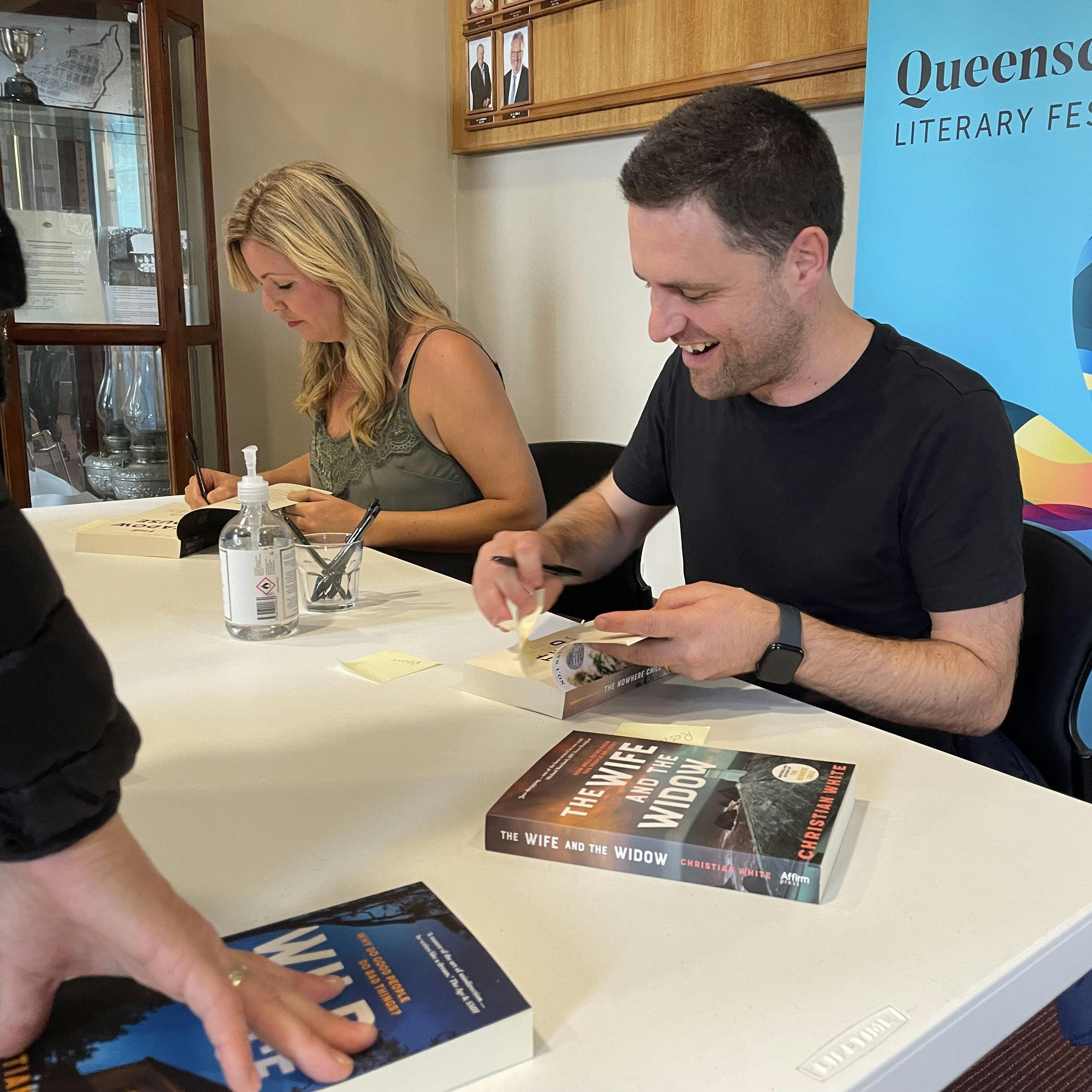 A woman and a man are seated beside eachother as they both sign copies of their books