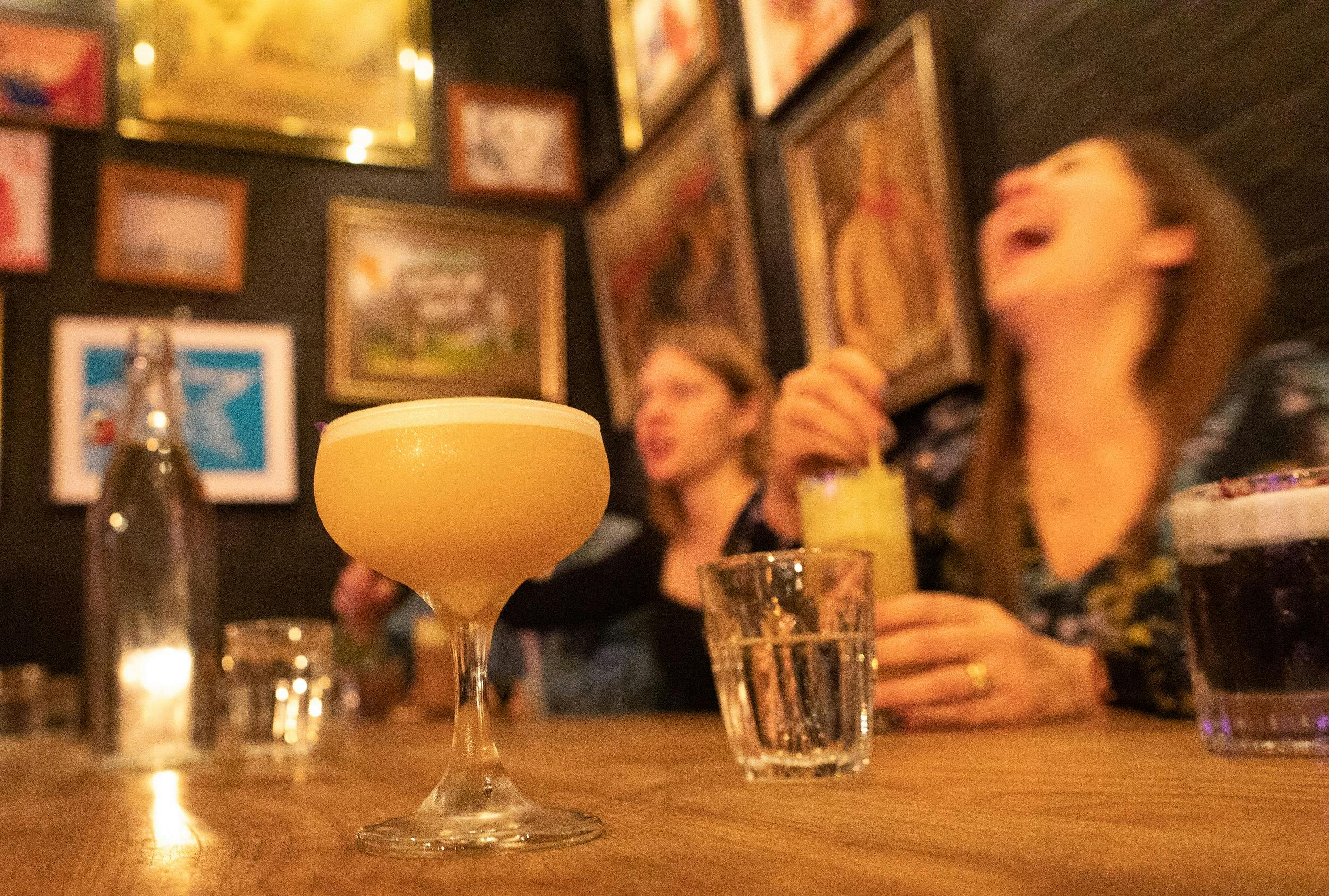 Two women sitting at a table drinking cocktails.  There is another cocktail in the foreground.