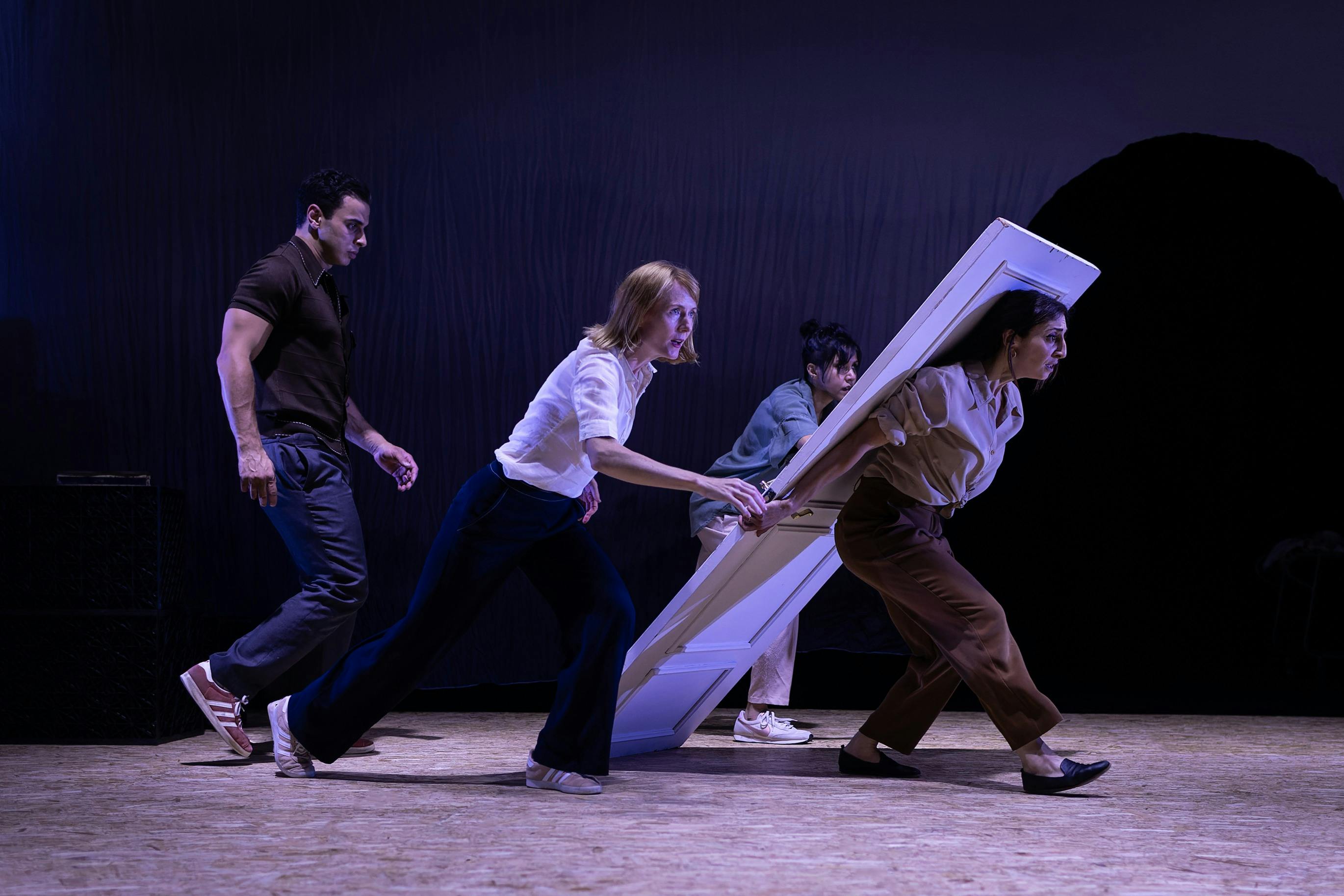 Four theater performers rush a large white door across a dim, purple-lit stage, creating a comic, ur