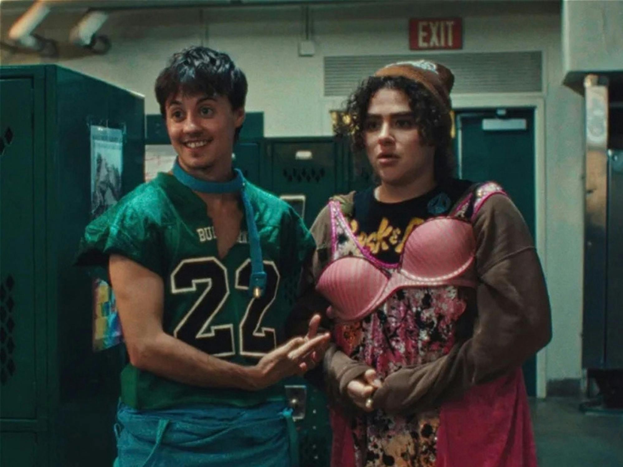 2 teenagers in a locker room, 1 wears a football jersey the other with bra over their clothes