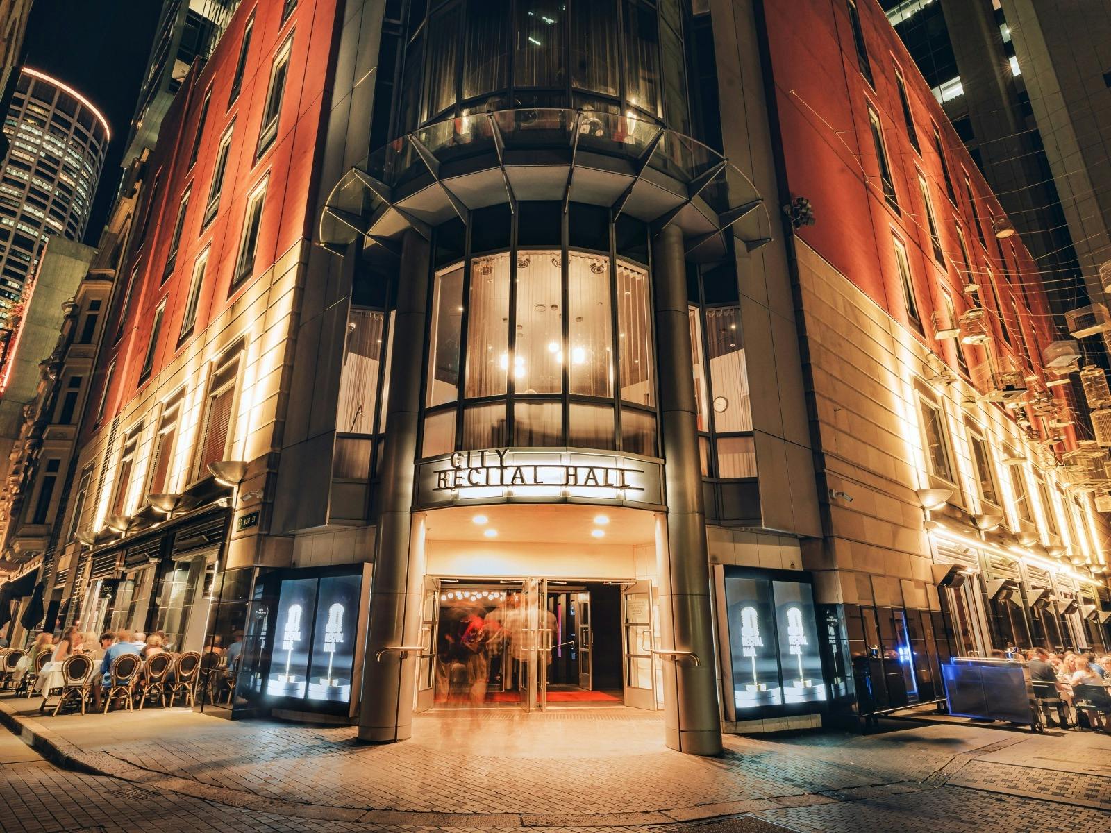 Night view of City Recital Hall entrance, illuminated facade with glass canopy and people entering,