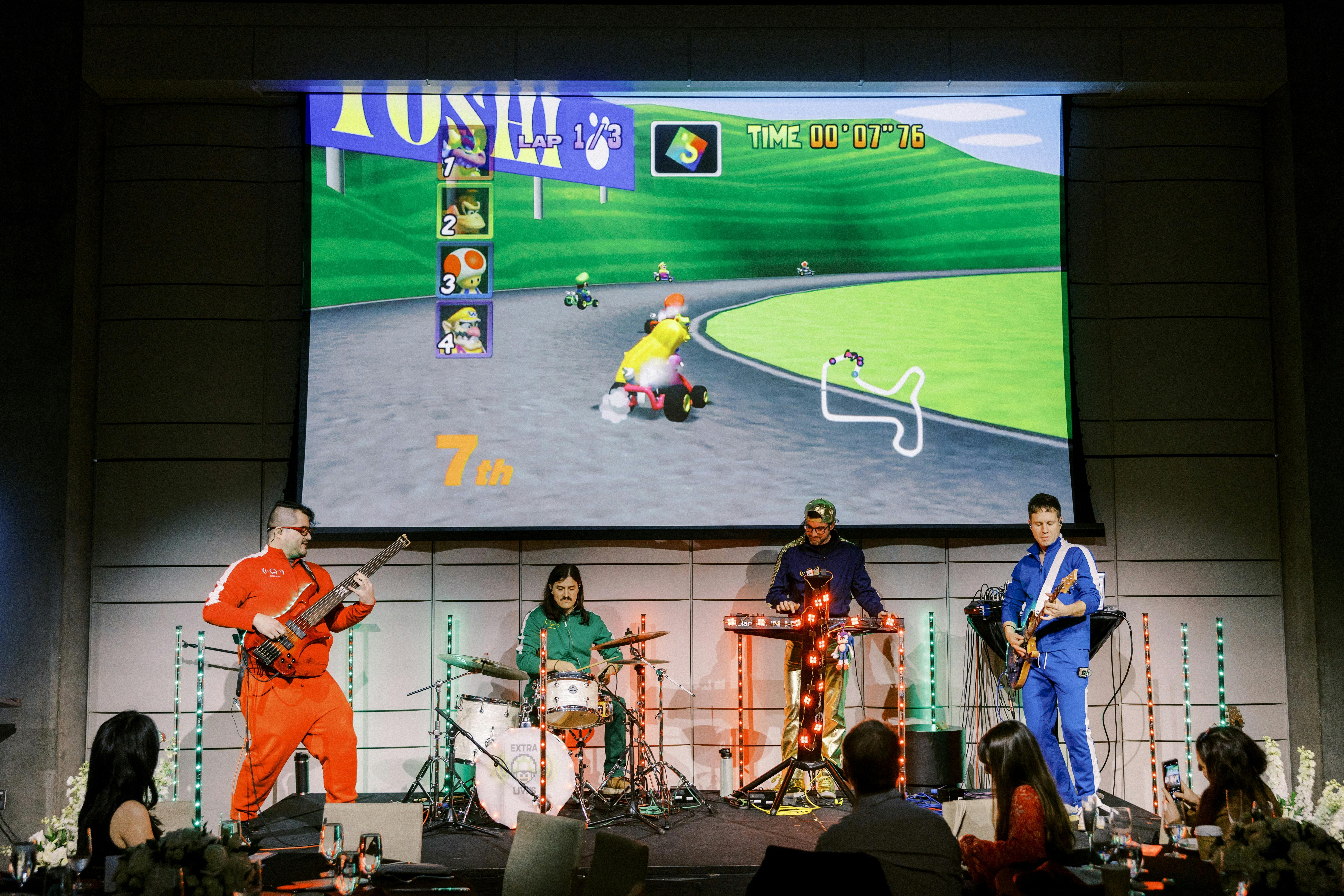 Live four piece band infront of a projector screen displaying the Game Mario Kart