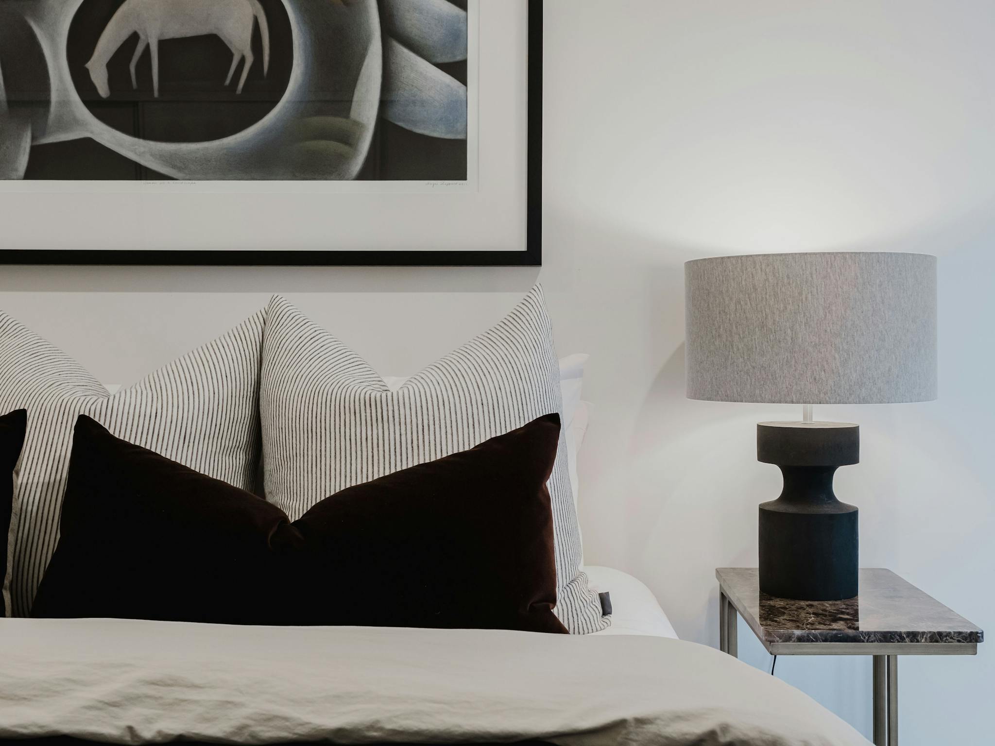 Bedroom with bed, pillows and cushions, tableside lamp and painting hanging on the wall